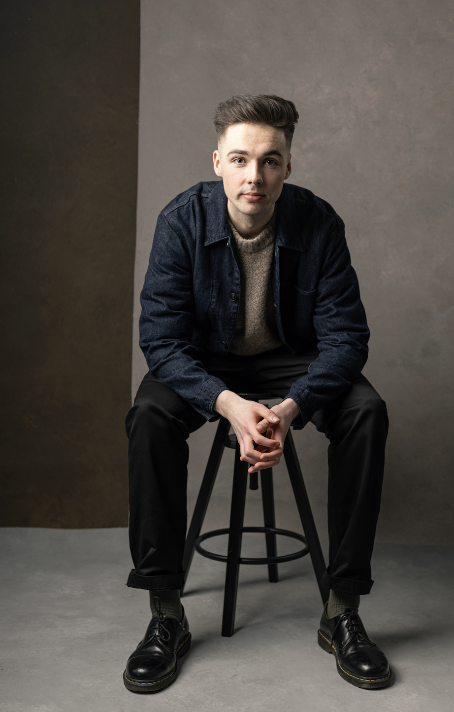A young man with short brown hair, wearing a denim jacket over a beige sweater, black pants, and black shoes, sitting on a stool against a neutral background.