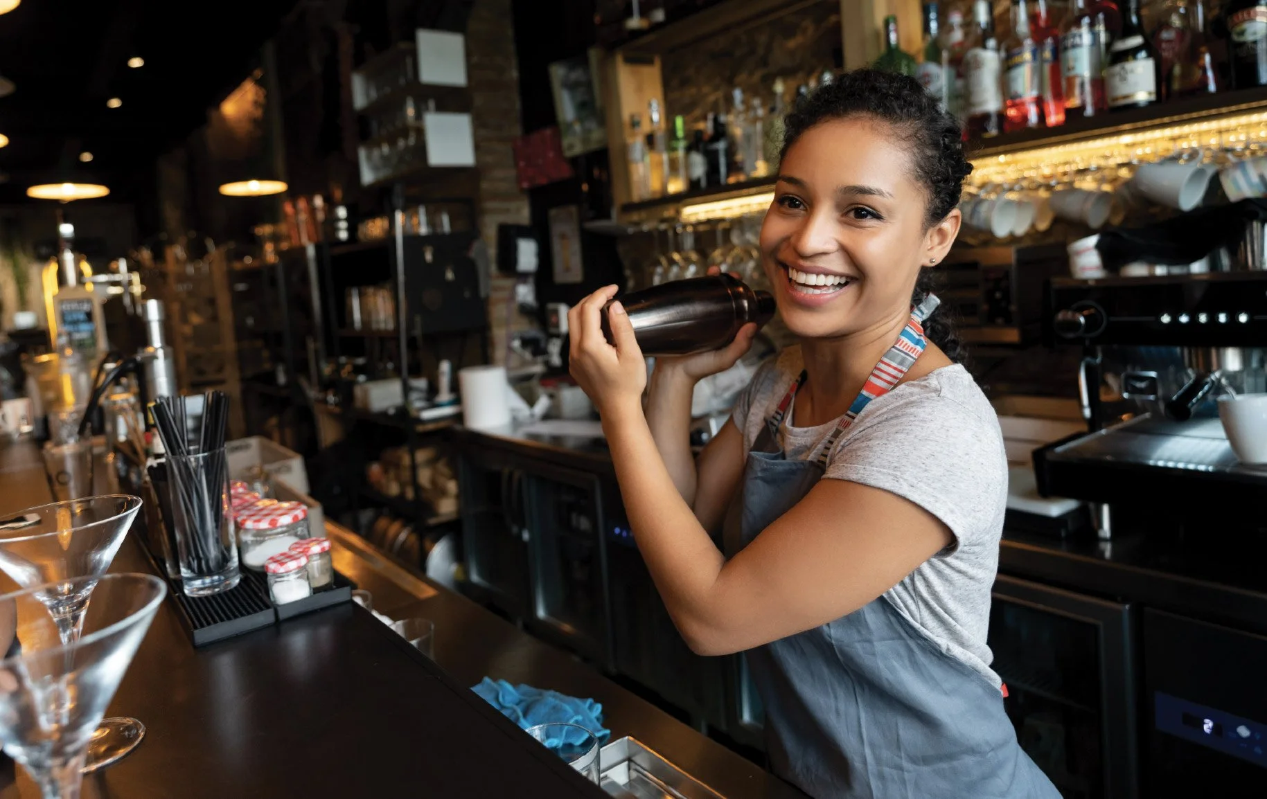A smiling female bartender with curly hair in a ponytail, wearing a gray t-shirt and apron, behind a bar counter in a cafe or bar. She is holding a dark bottle near her cheek and appears to be laughing or talking.