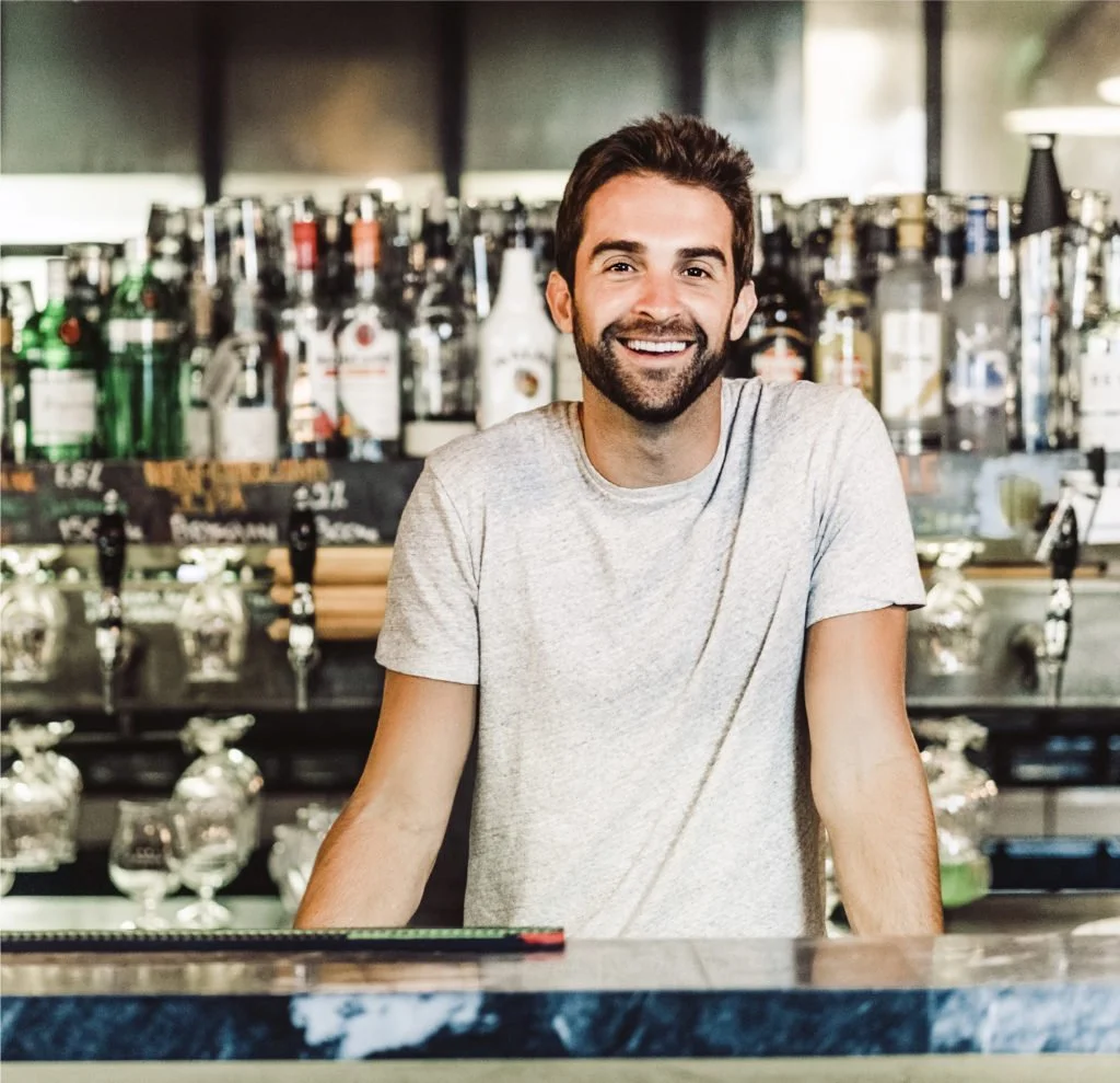 Smiling man with brown hair and beard standing at bar with bottles of alcohol behind him.