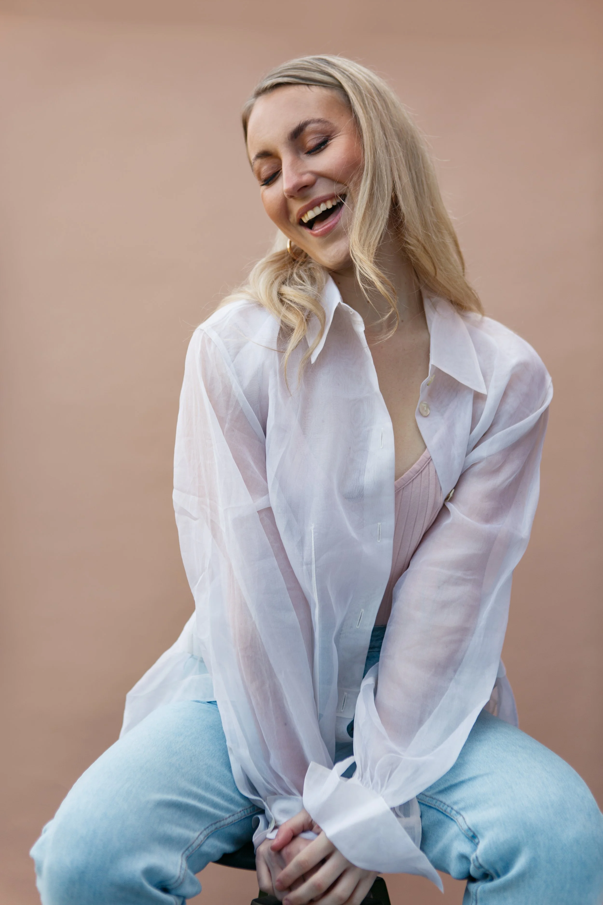 A young woman with blonde hair sitting cross-legged and laughing, wearing a white button-up shirt and light blue jeans against a plain, light brown background.