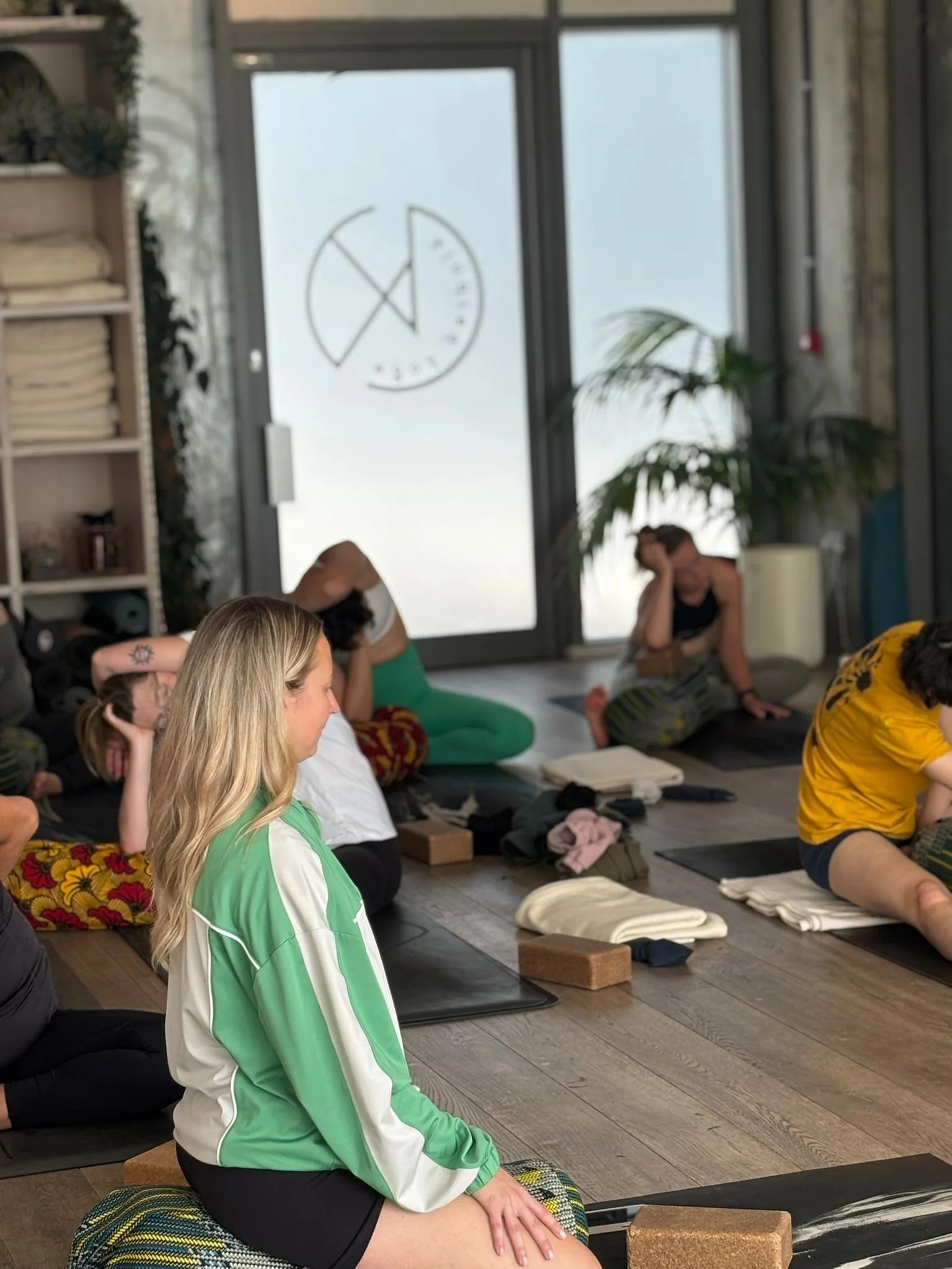 People sitting cross-legged on yoga mats in a serene indoor space, practicing meditation or mindfulness, with a large window and houseplants in the background.