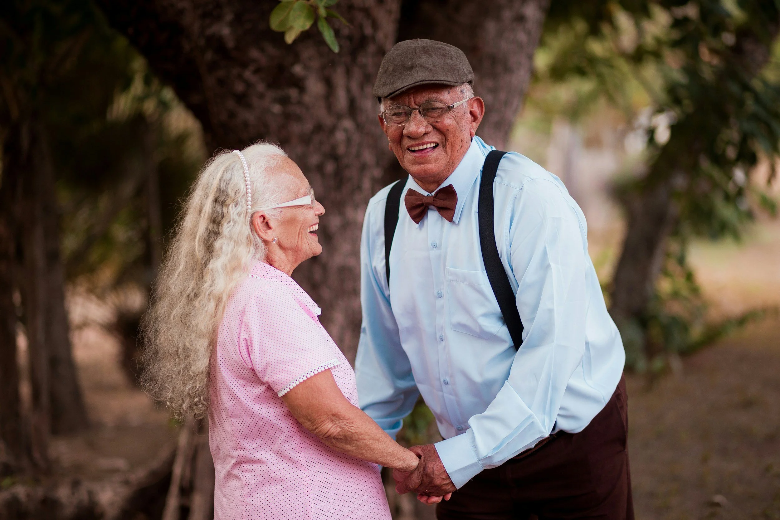An elderly couple holding hands and smiling at each other outdoors near a tree.