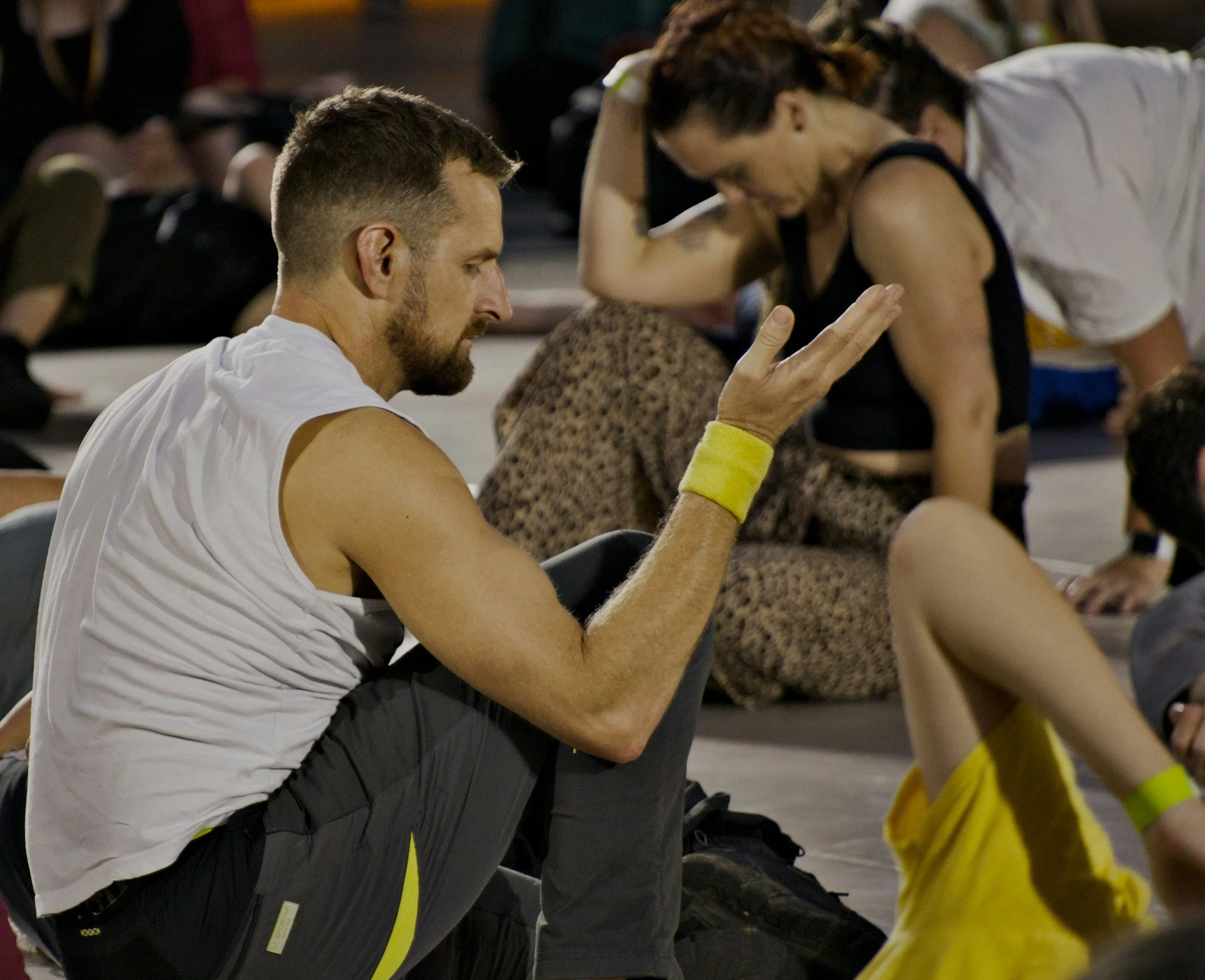 A man with a beard, wearing a white sleeveless shirt and yellow wristband, sits on the floor with his eyes closed and hands raised in a meditative or prayerful gesture. There are women in the background, also engaged in the same activity, in a group setting that appears to be a yoga or meditation class.