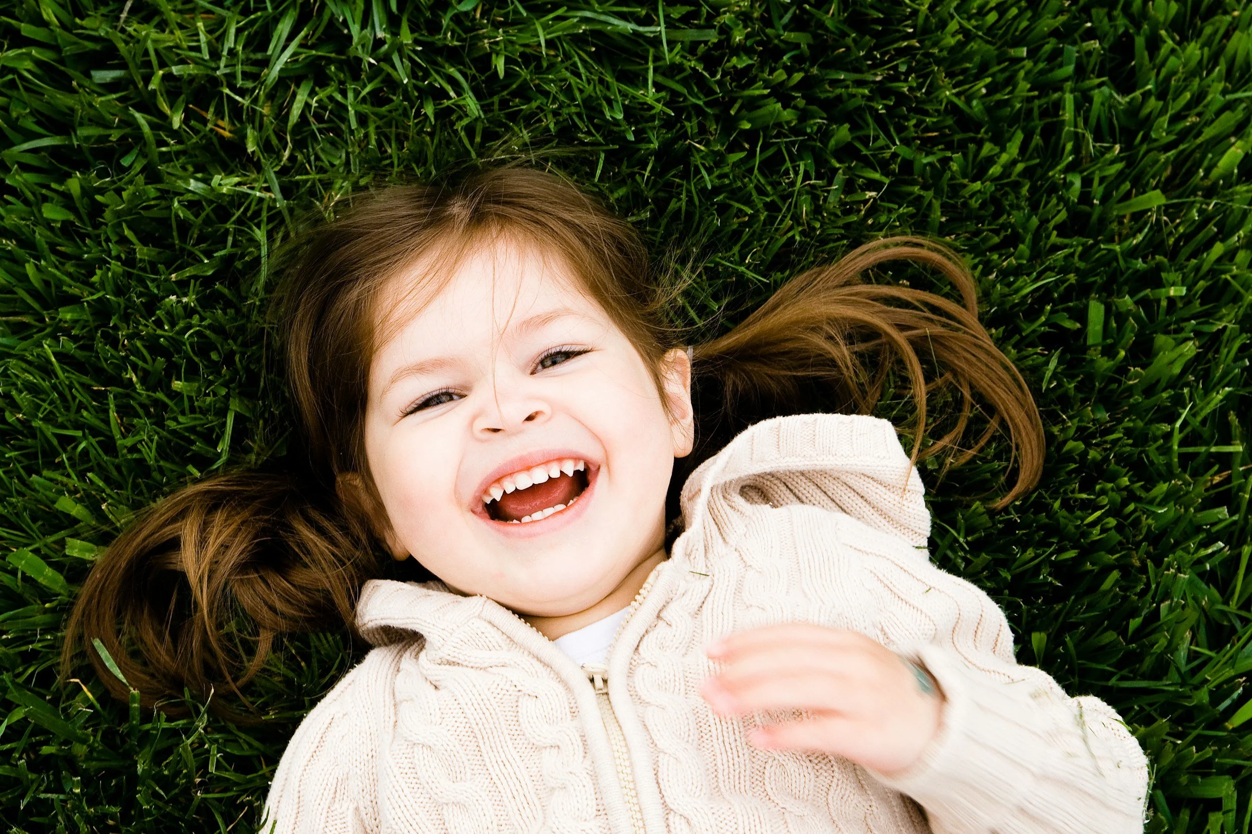 A young girl with long brown hair lying on green grass, smiling and laughing.