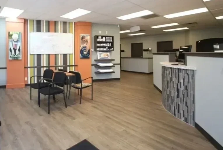 A waiting area with four black chairs and a small table, colorful striped walls, a whiteboard, and shelves on the wall in a modern office or clinic reception area.