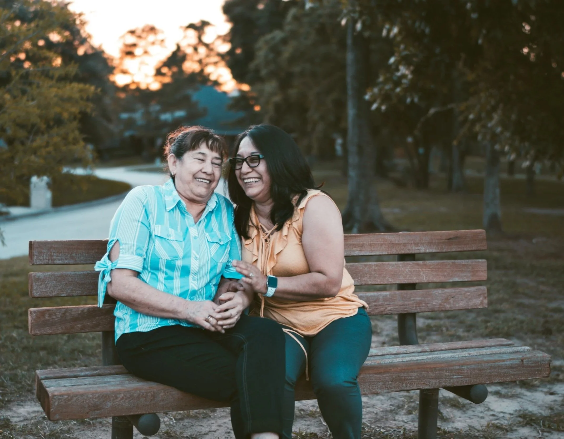 Two women sitting on a park bench, laughing and enjoying each other's company during sunset, with trees and a pathway in the background.