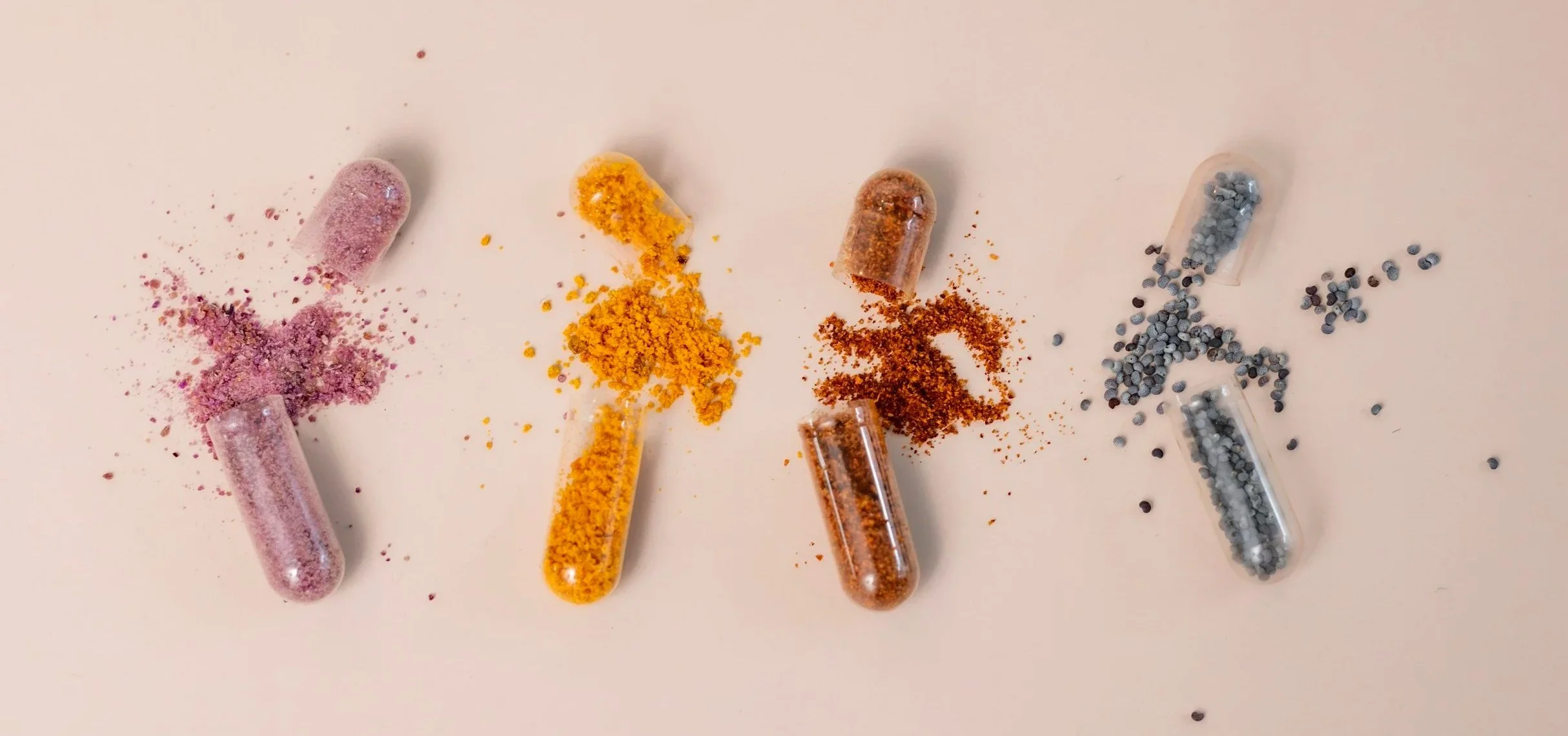 Four capsules spilling colored powders in pink, yellow, brown, and gray on a white surface.