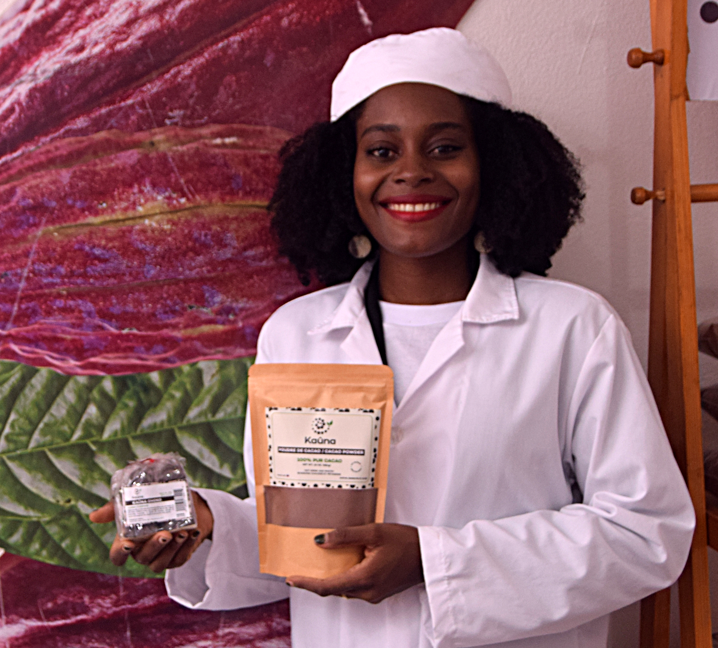 A woman with dark curly hair and dark skin, smiling and wearing a white coat and white cap, holding a package of cacao and a small item, standing in front of a colorful plant backdrop.