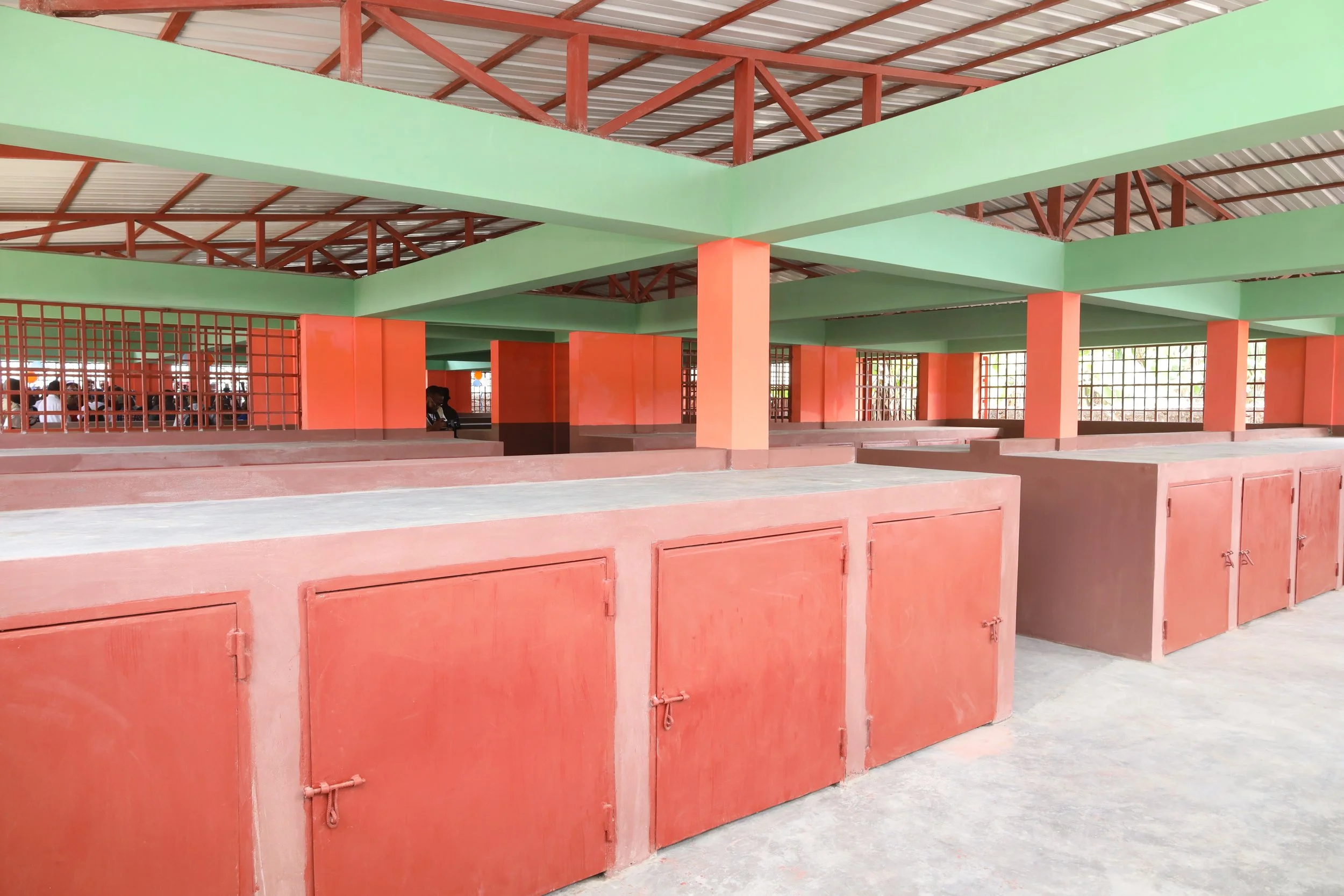 Indoor space with reddish-pink storage cabinets, green beams, metal roof, and open grid windows, with a person visible in the background.