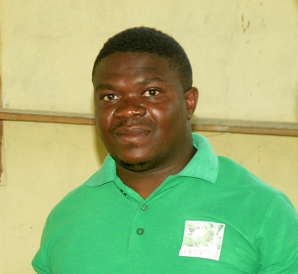 A man wearing a green polo shirt with a white patch on it, standing in front of a beige wall.