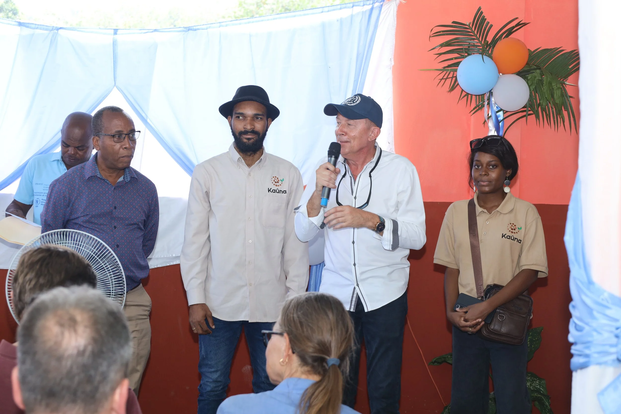 Group of people standing on a stage, with one man speaking into a microphone, at an indoor event decorated with blue and orange balloons, green leaves, and light blue curtains.