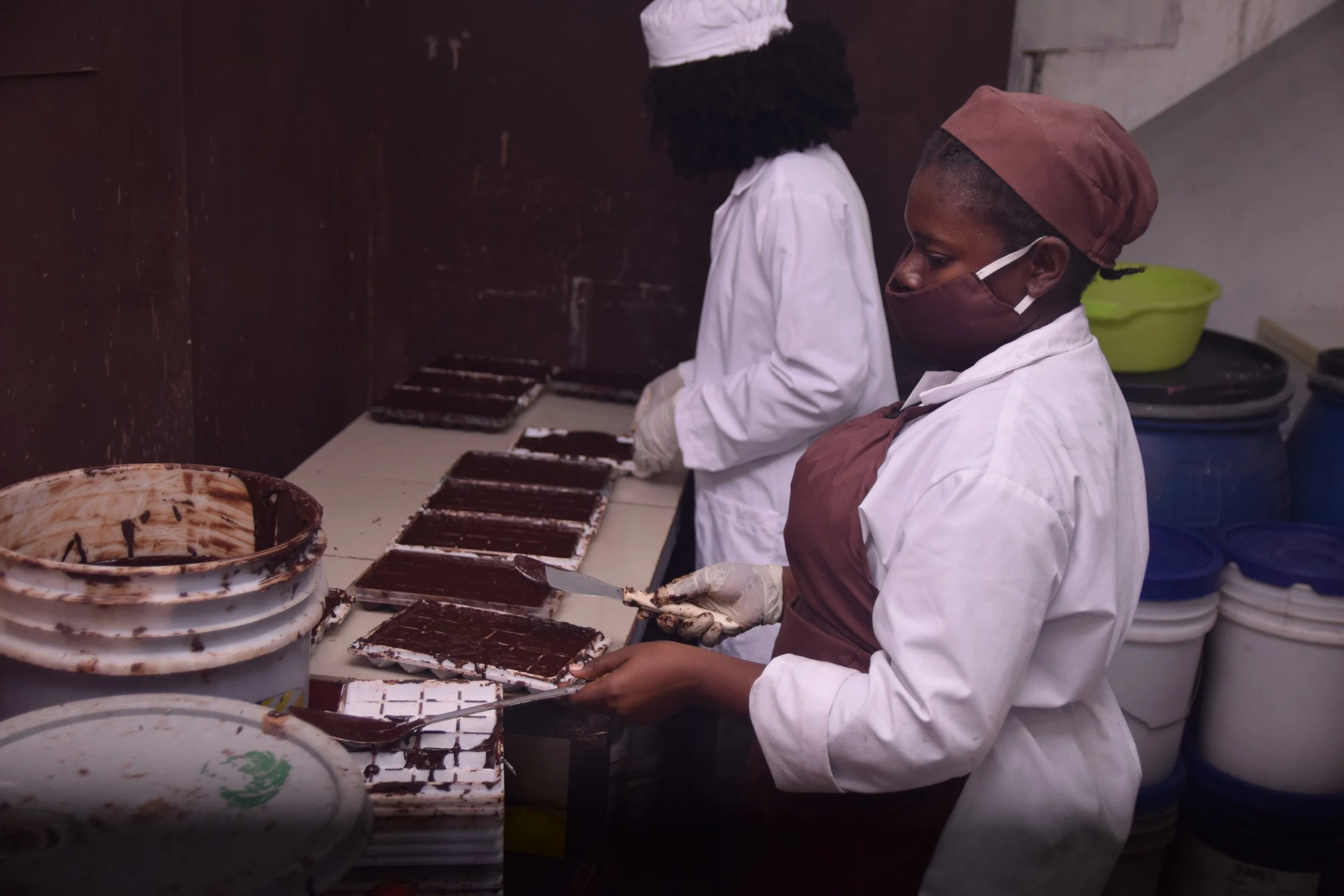 Two people in chef uniforms and face masks preparing or packaging chocolate bars in a kitchen.