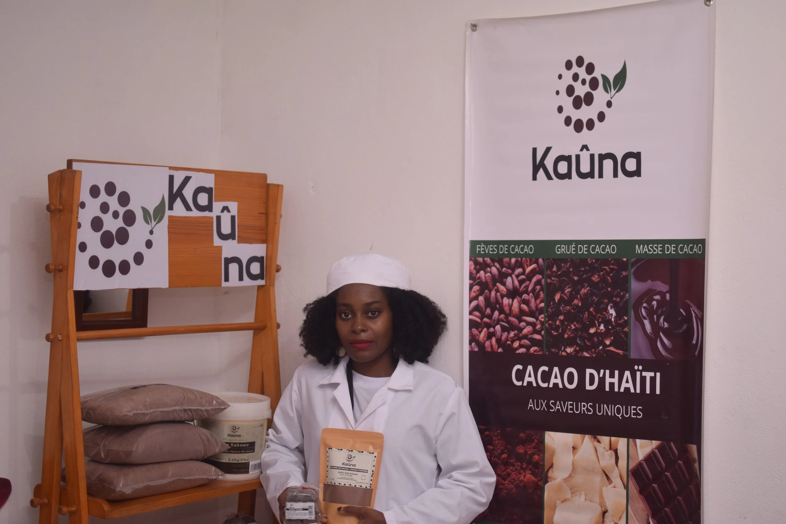 Woman standing in front of a Kissimmee, Hawaii, with a white coat and a hat, holding a package of Haiti cacao, with a large poster behind her displaying images of cacao and chocolate, and a small wooden display shelf with pre-packaged cacao products 