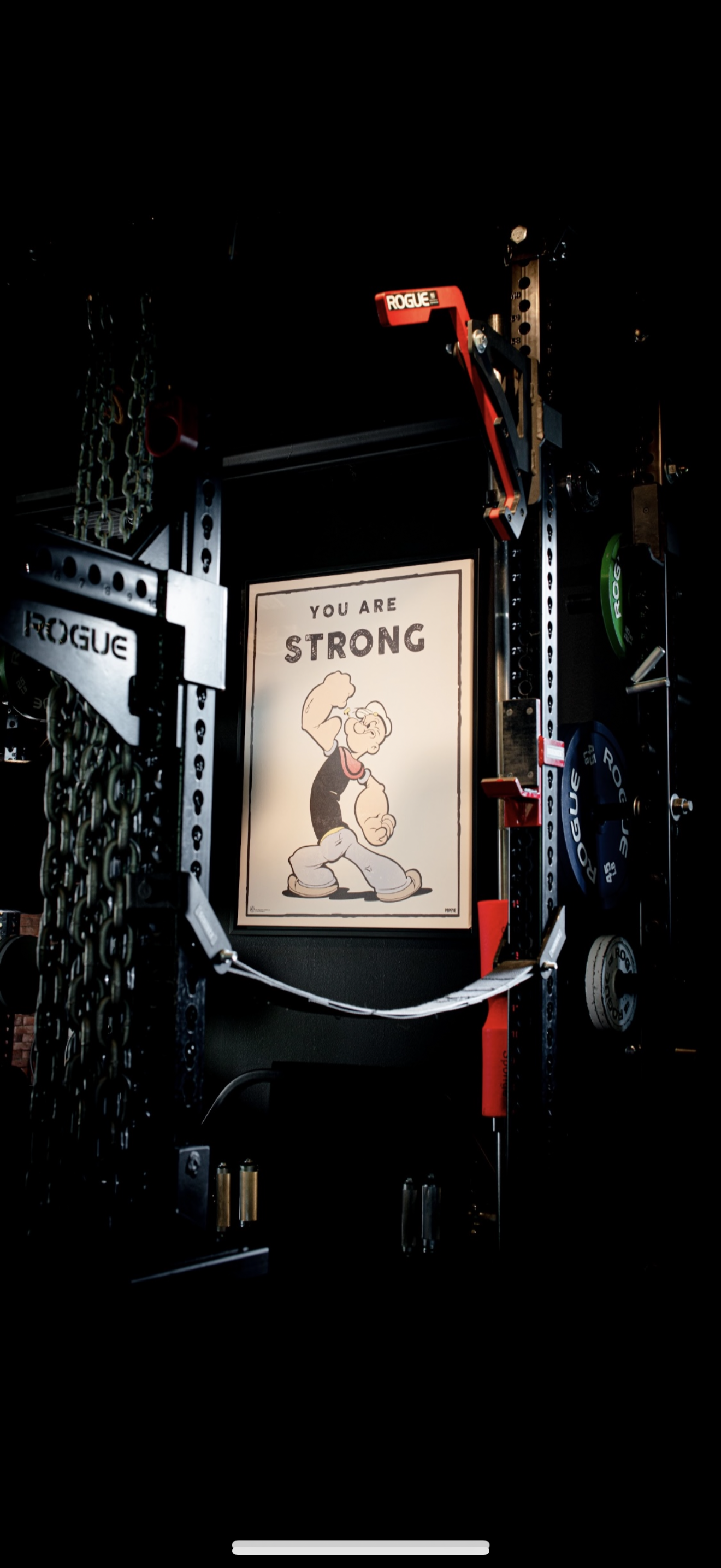 A gym wall with inspirational poster saying 'You Are Strong' and featuring a muscular cartoon character. Surrounding it are various gym equipment such as chains, weights, and a red rack with the brand name Rogue.