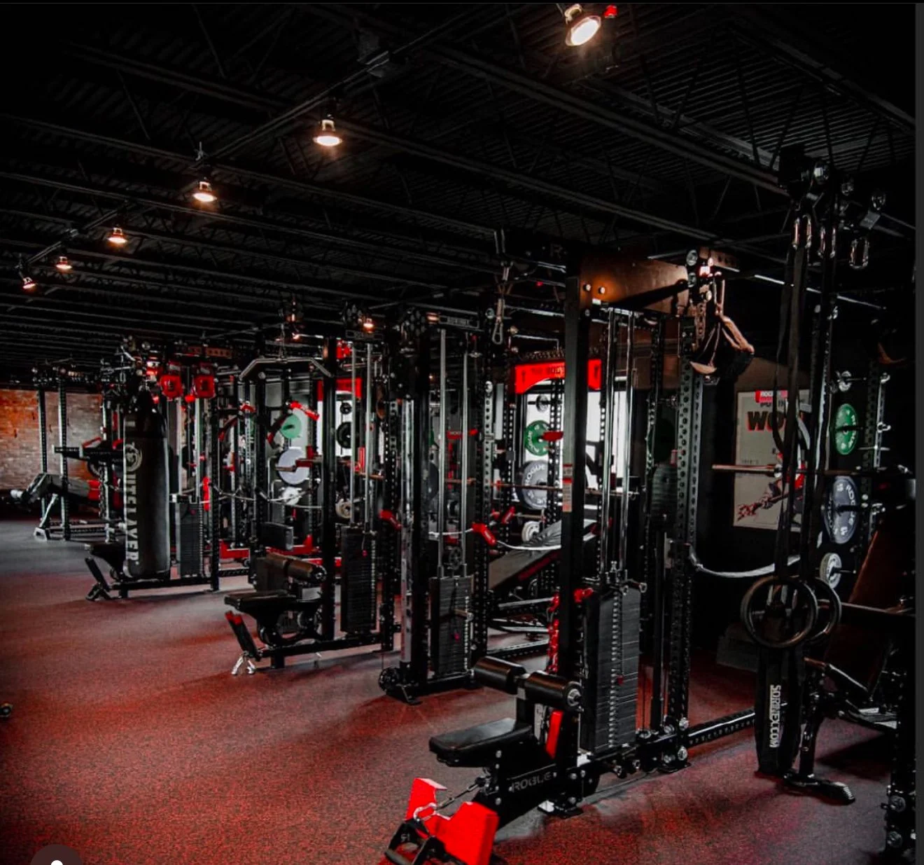 An interior view of a gym with various exercise equipment including weight machines, barbells, and rings, with dim lighting and exposed ceiling