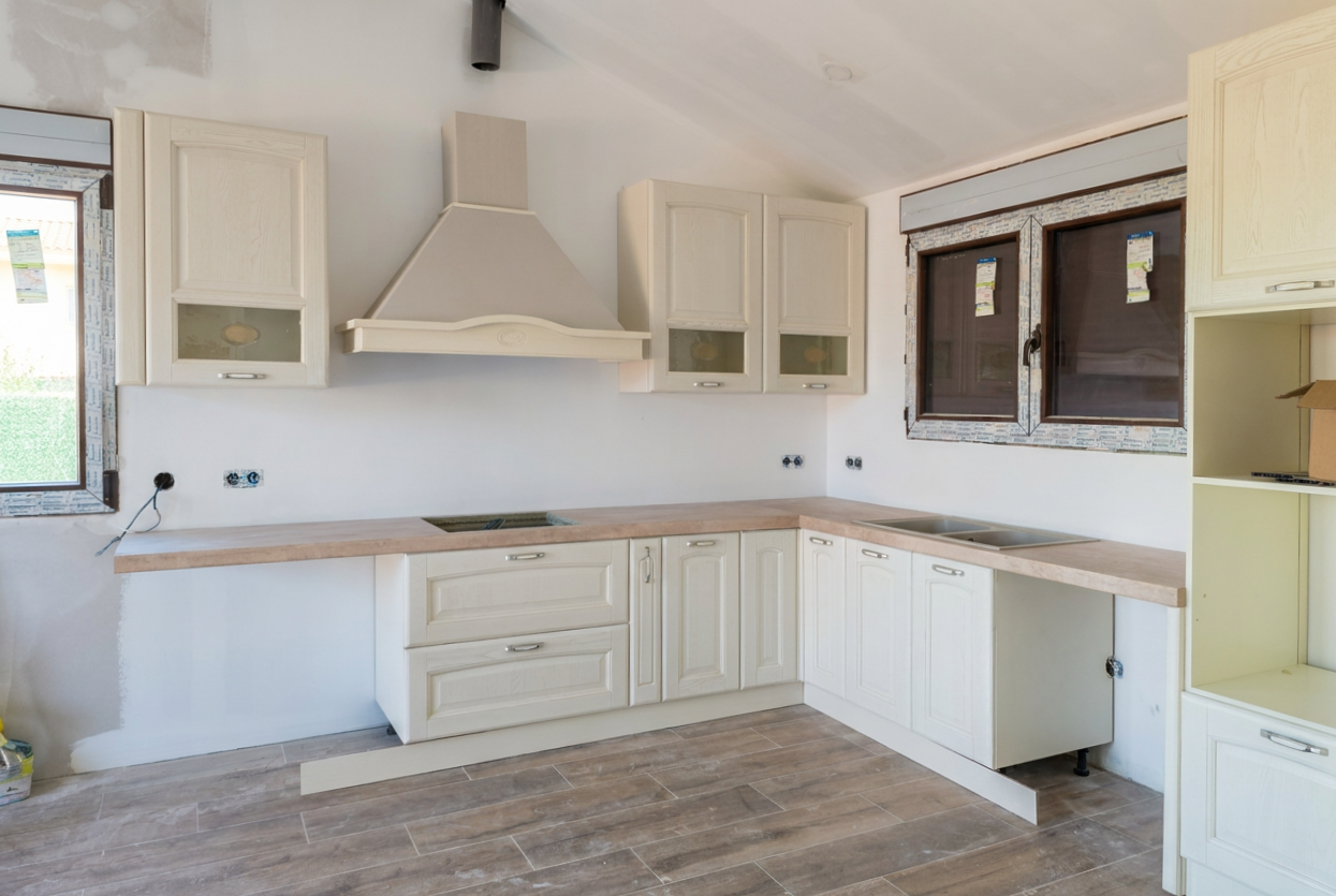 Cocina en construcción con gabinetes blancos, encimera de madera, ventanas con persianas, y un área para fregadero sin instalar.