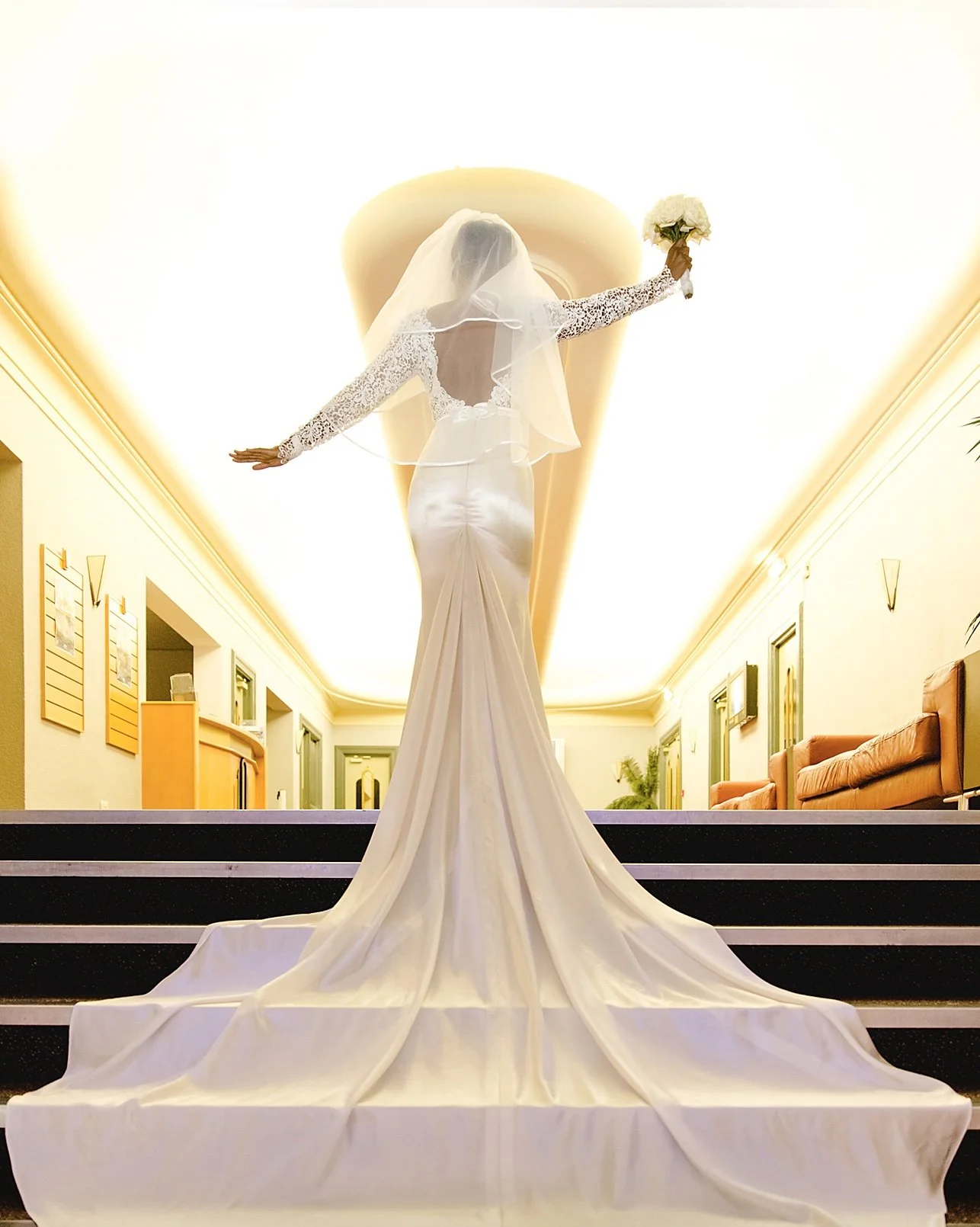 A bride in a white wedding gown with long train and lace sleeves, holding a bouquet of white flowers, standing on a staircase in an elegant, well-lit indoor hall.