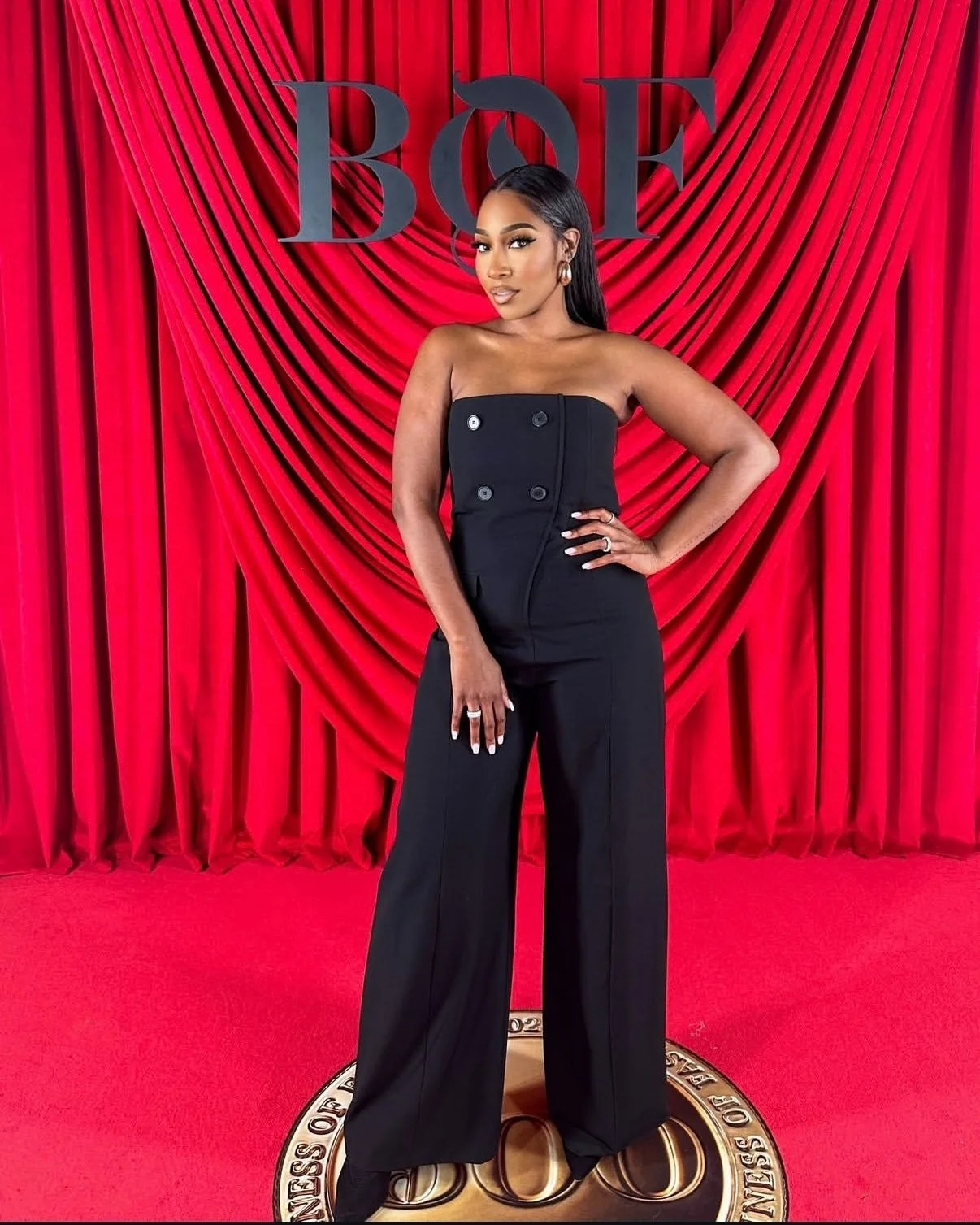 A woman in a black strapless jumpsuit posing on a red carpet in front of a red curtain with the BOSS logo.