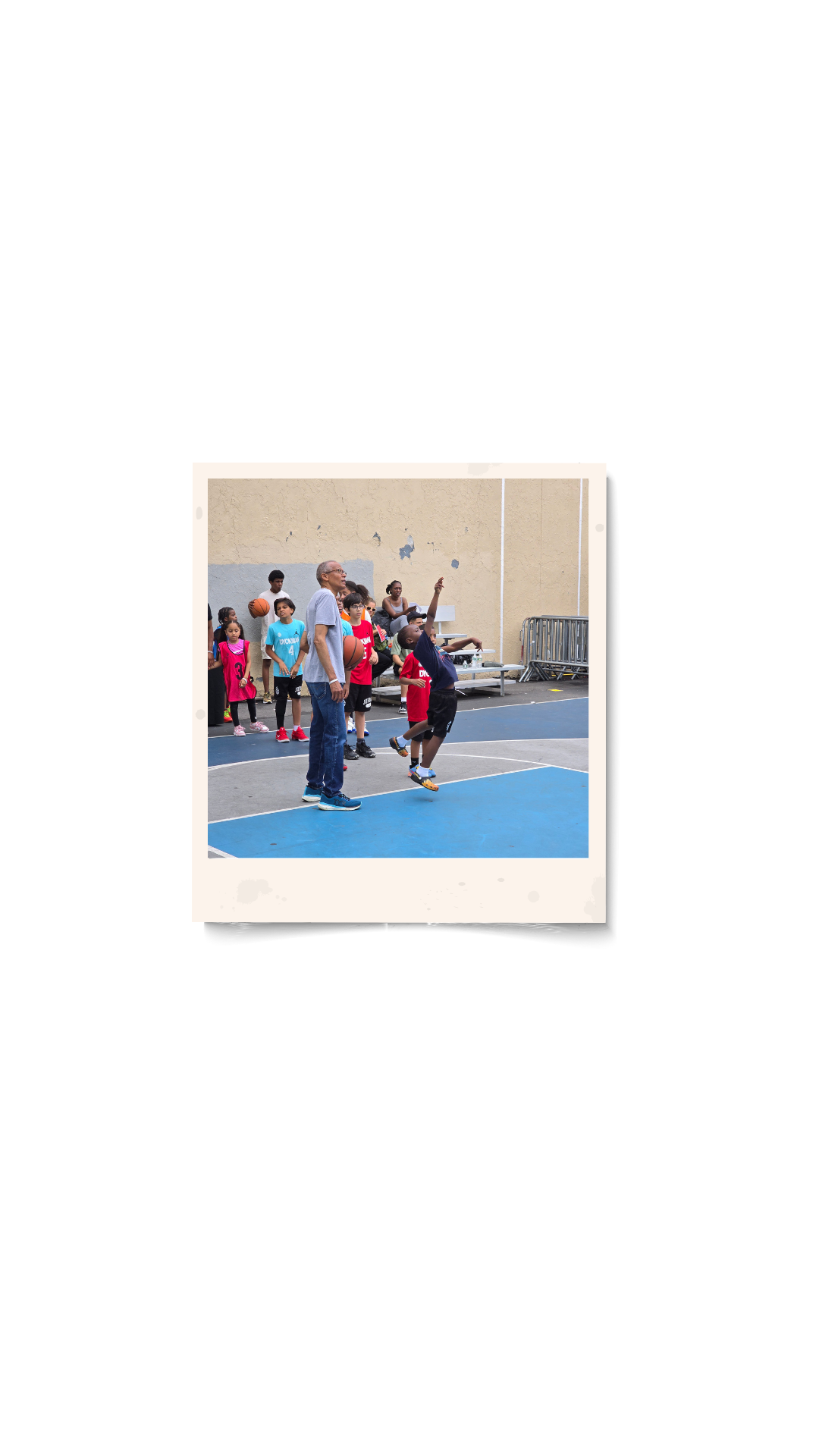 Kids playing basketball on an outdoor court with an adult coach and a group of children watching.
