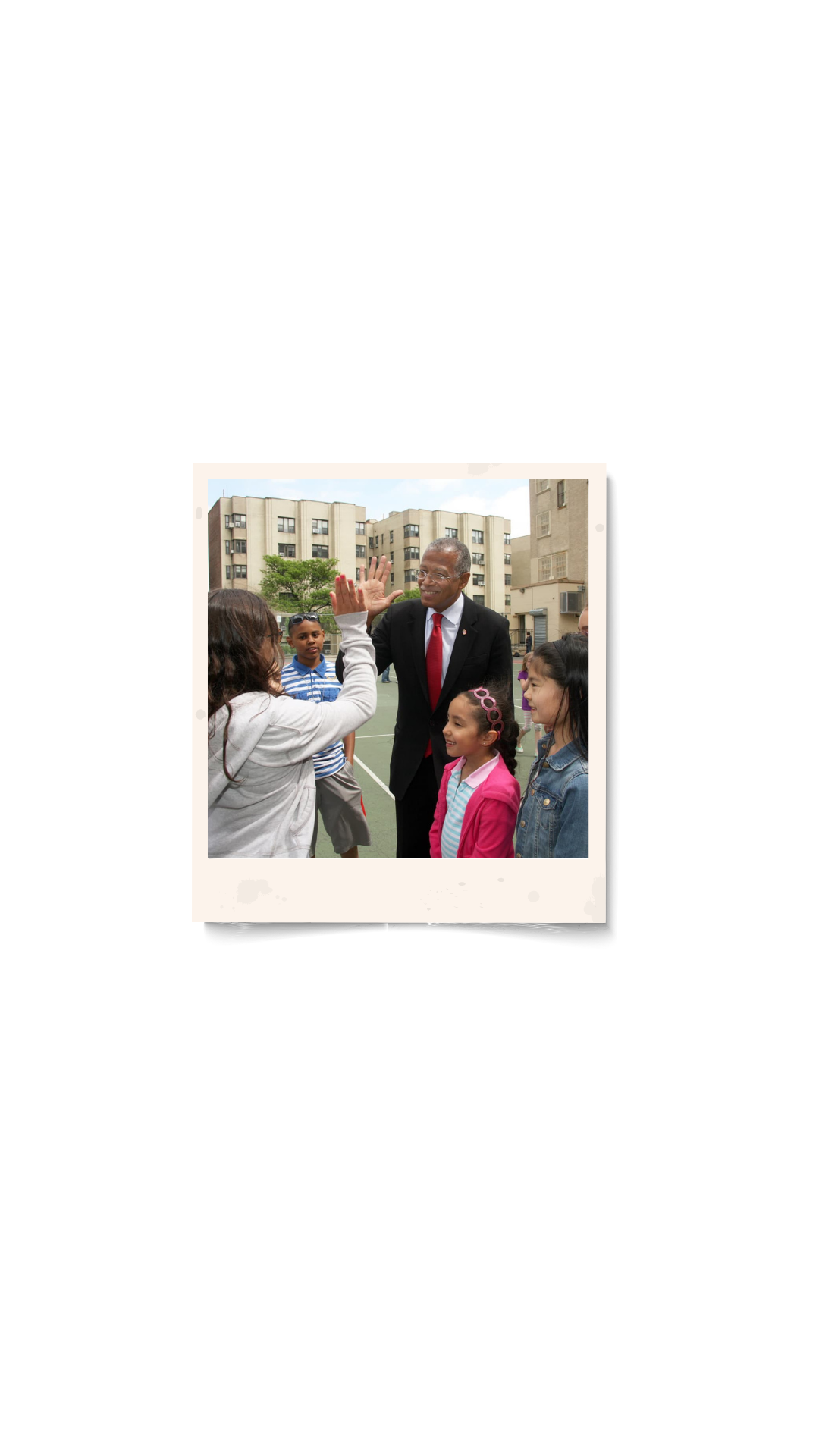 An outdoor gathering of children and an adult man in a suit giving a high-five on a playground, with residential buildings in the background.