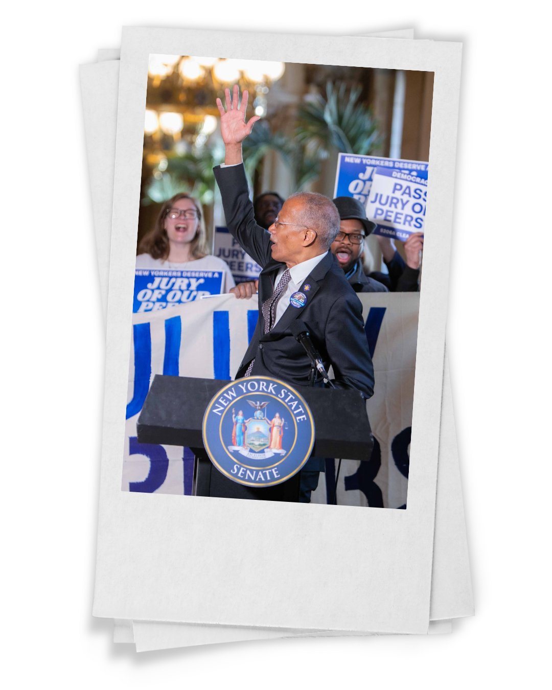 A man speaking at a New York State Senate podium with a crowd of people behind him holding signs that read 'Jury of Our Peers' and 'New Yorkers Deserve'. The man is wearing glasses, a suit, and a tie, and is waving to the crowd.
