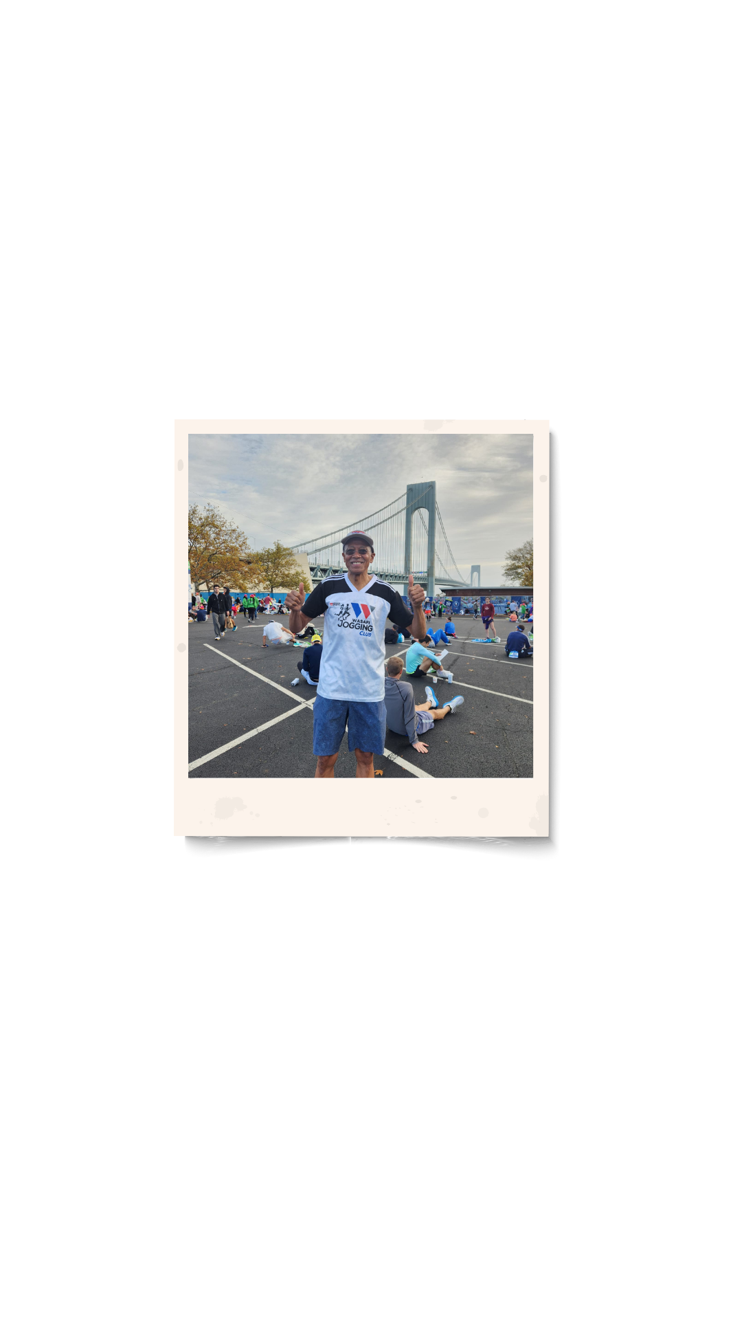 Person smiling and giving thumbs-up at an outdoor event near a large bridge, with other people sitting on the ground in the background.