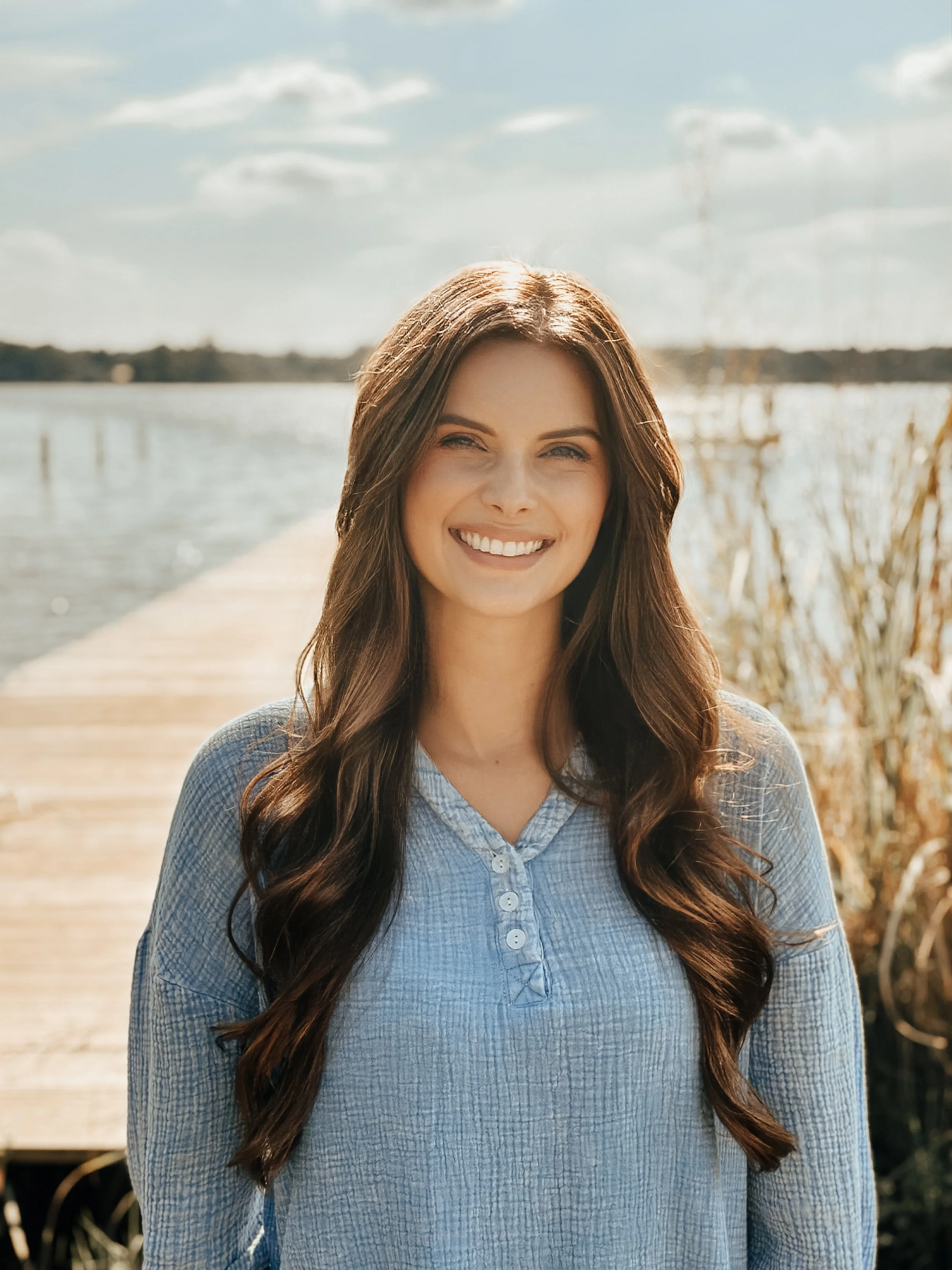 A smiling woman with long brown hair standing on a wooden dock by a lake on a sunny day.