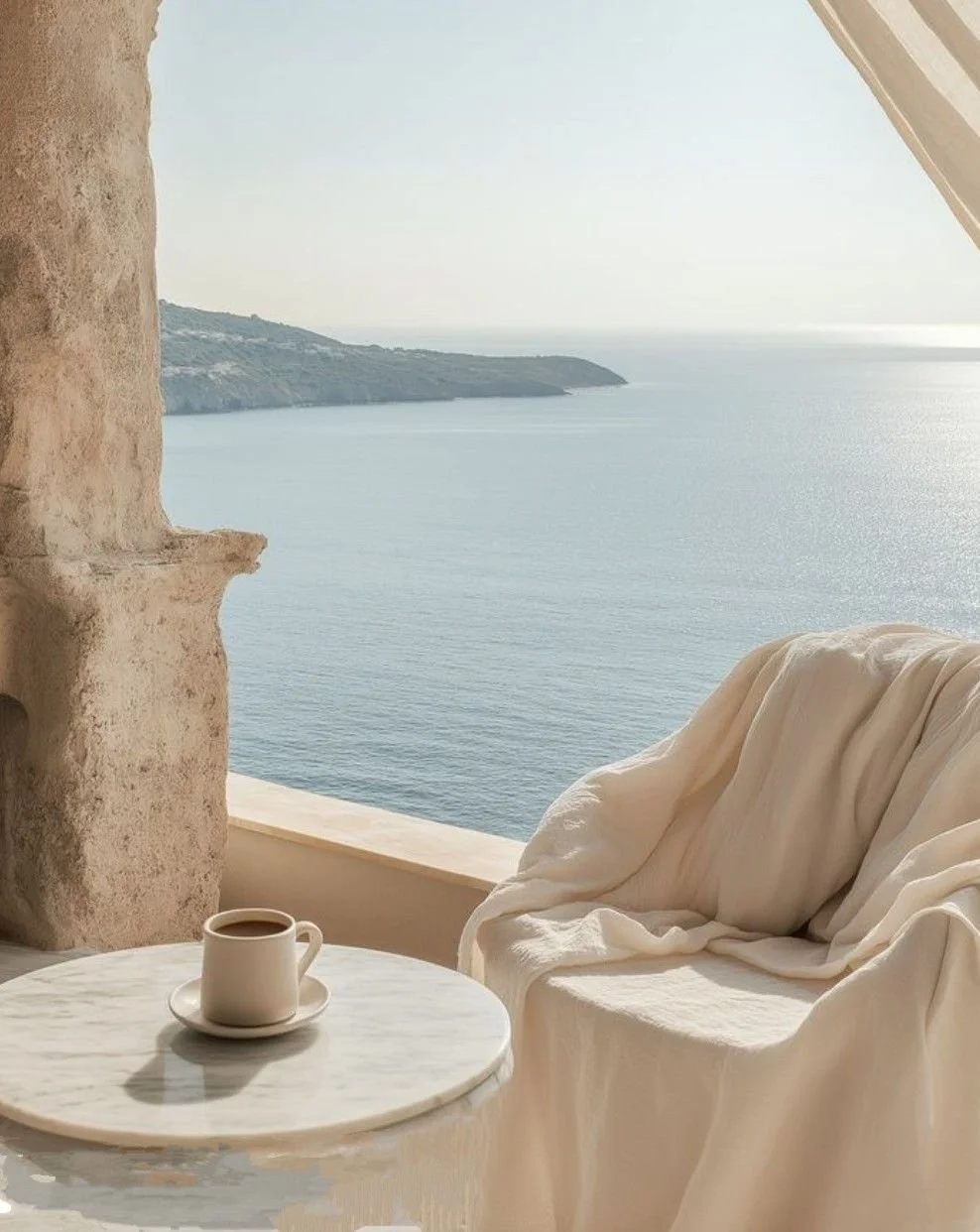 A cozy seating area with a white draped sofa and a marble side table holding a cup of coffee, overlooking a serene ocean view through an open window with stone architecture.