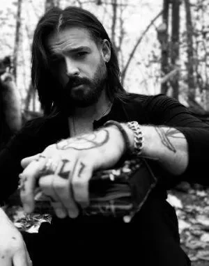 A man with long hair and a beard in a forest setting, holding a camera and looking at the viewer.