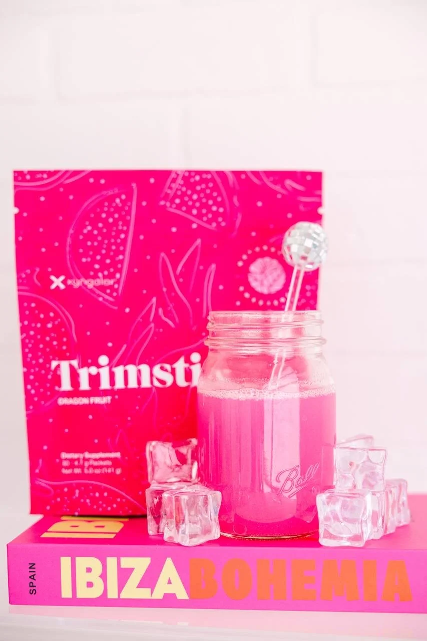 A pink box of Trimsti dragon fruit dietary supplement set against a pink and white background, with a pink glass jar filled with pink liquid, topped with a decorative stick with a small mirrored ball, and surrounded by ice cubes, all placed on a pink "Ibiza Bohemia" book.