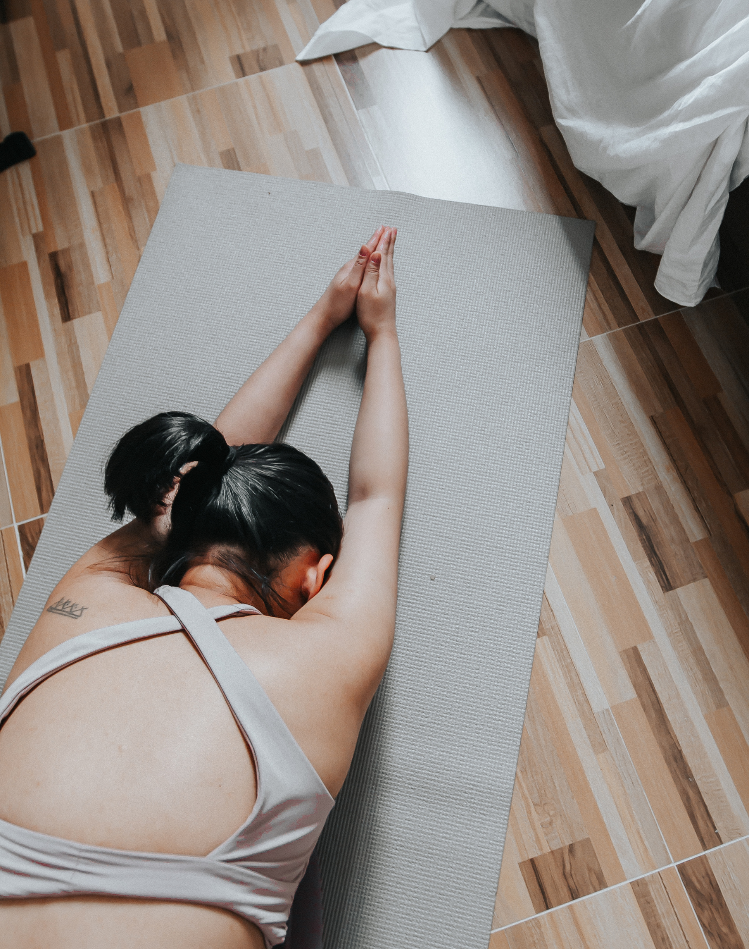 A woman practicing yoga on a grey mat in a room with wooden floor. She has dark hair tied in a ponytail and is wearing a white sports bra. Her head is resting on her folded arms.