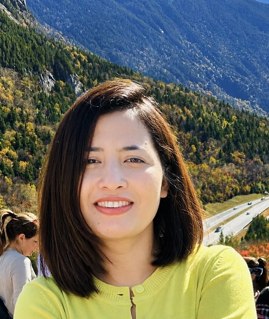 A woman with shoulder-length dark hair wearing a yellow top, smiling outdoors with a mountain and forest backdrop and a highway below.