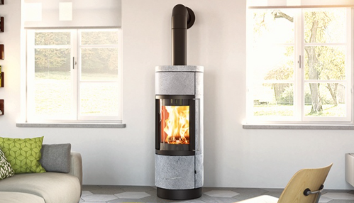 A modern living room with a wood-burning stove in the center, emitting flames. Two large windows on either side allow natural light to enter, and a gray sofa with green and gray pillows is on the left. A wooden chair is on the right.