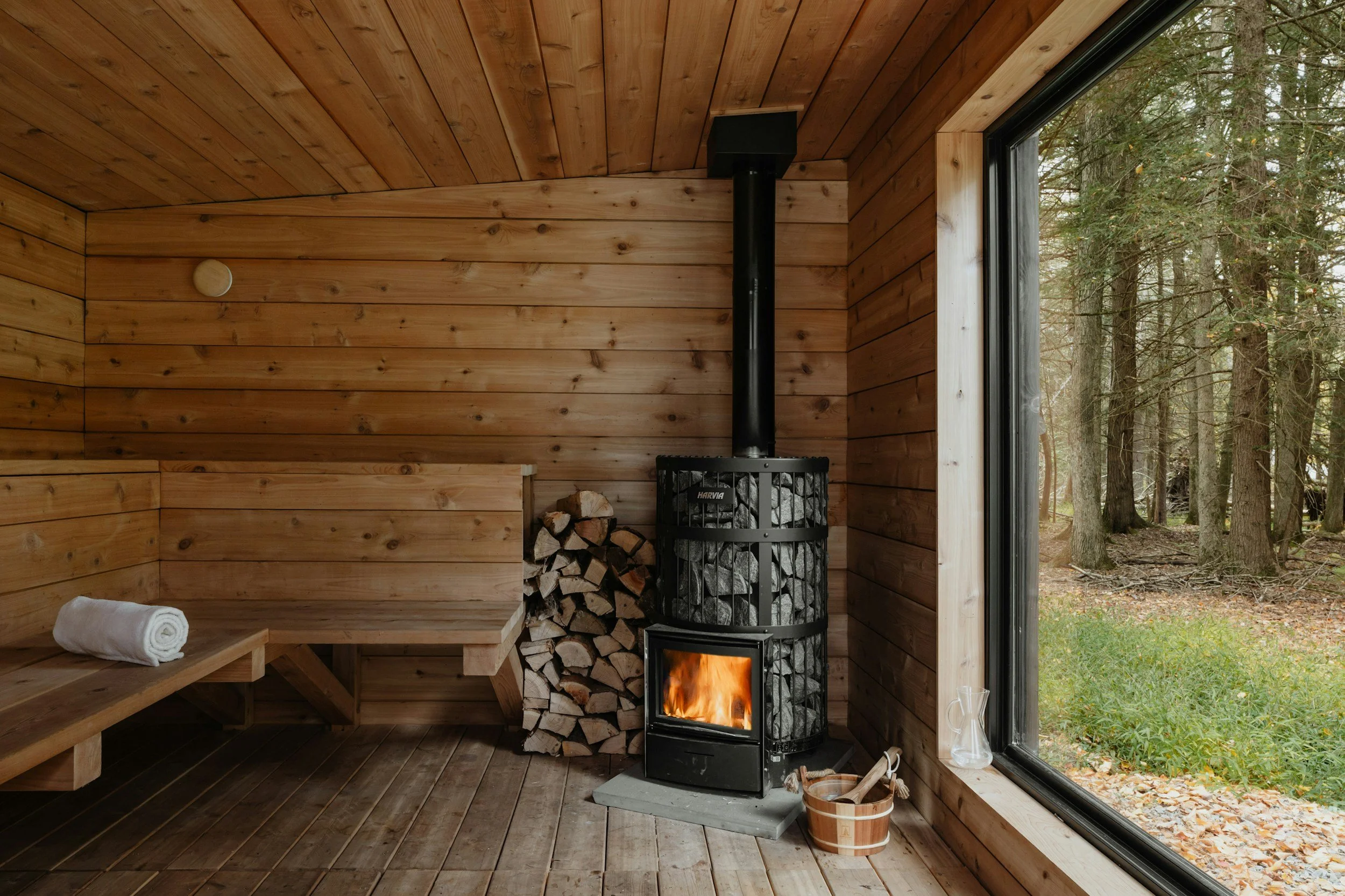 Interior of a wooden sauna with a wood-burning stove, stacked firewood, a bench with a rolled towel, and a large window showing a forest outside.