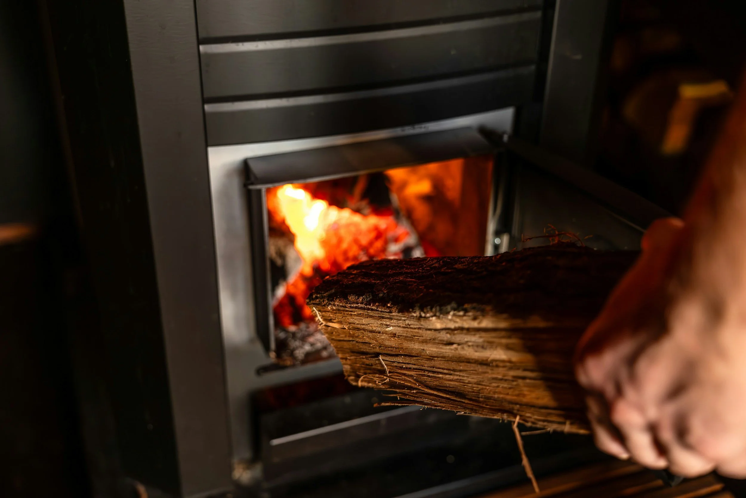 A person placing a log into a wood-burning stove with glowing flames inside.