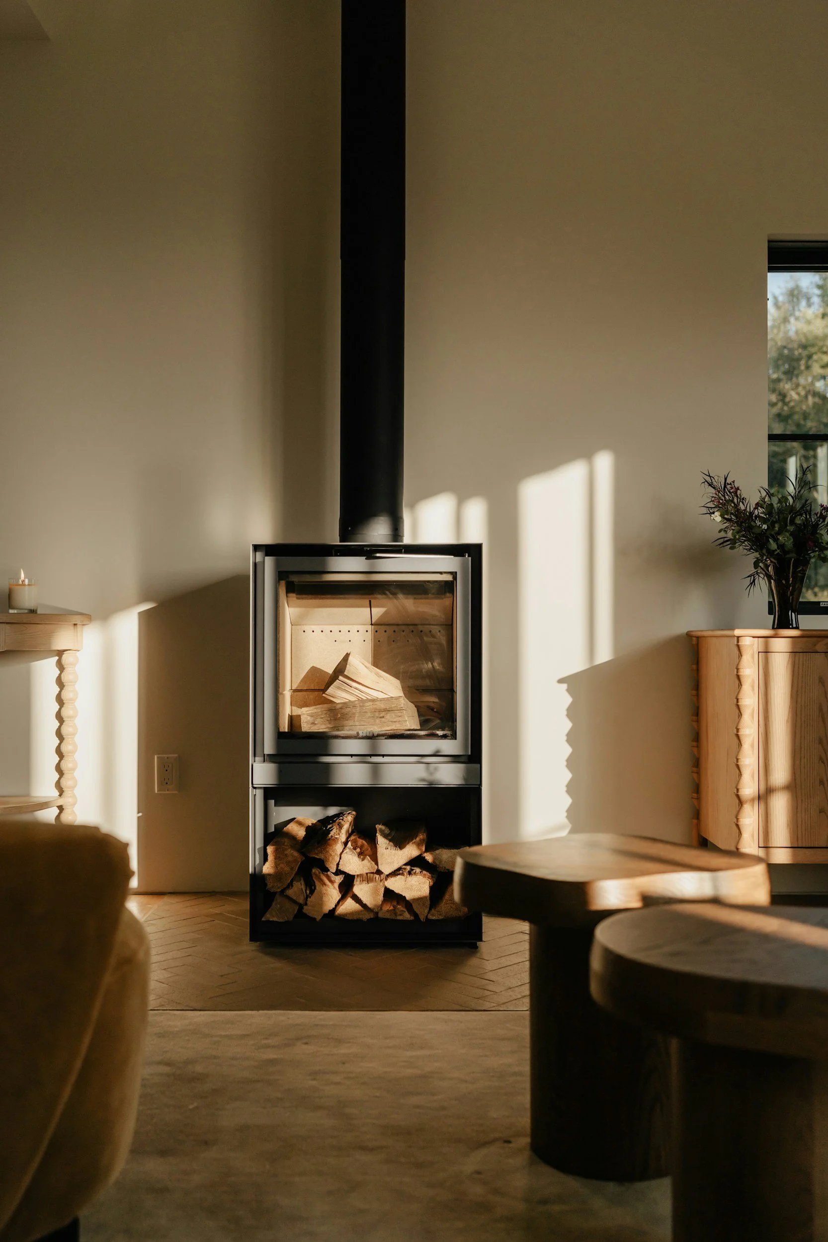 A modern indoor fireplace with stacked firewood below and flames inside, surrounded by minimalist wooden furniture, bathed in warm sunlight.