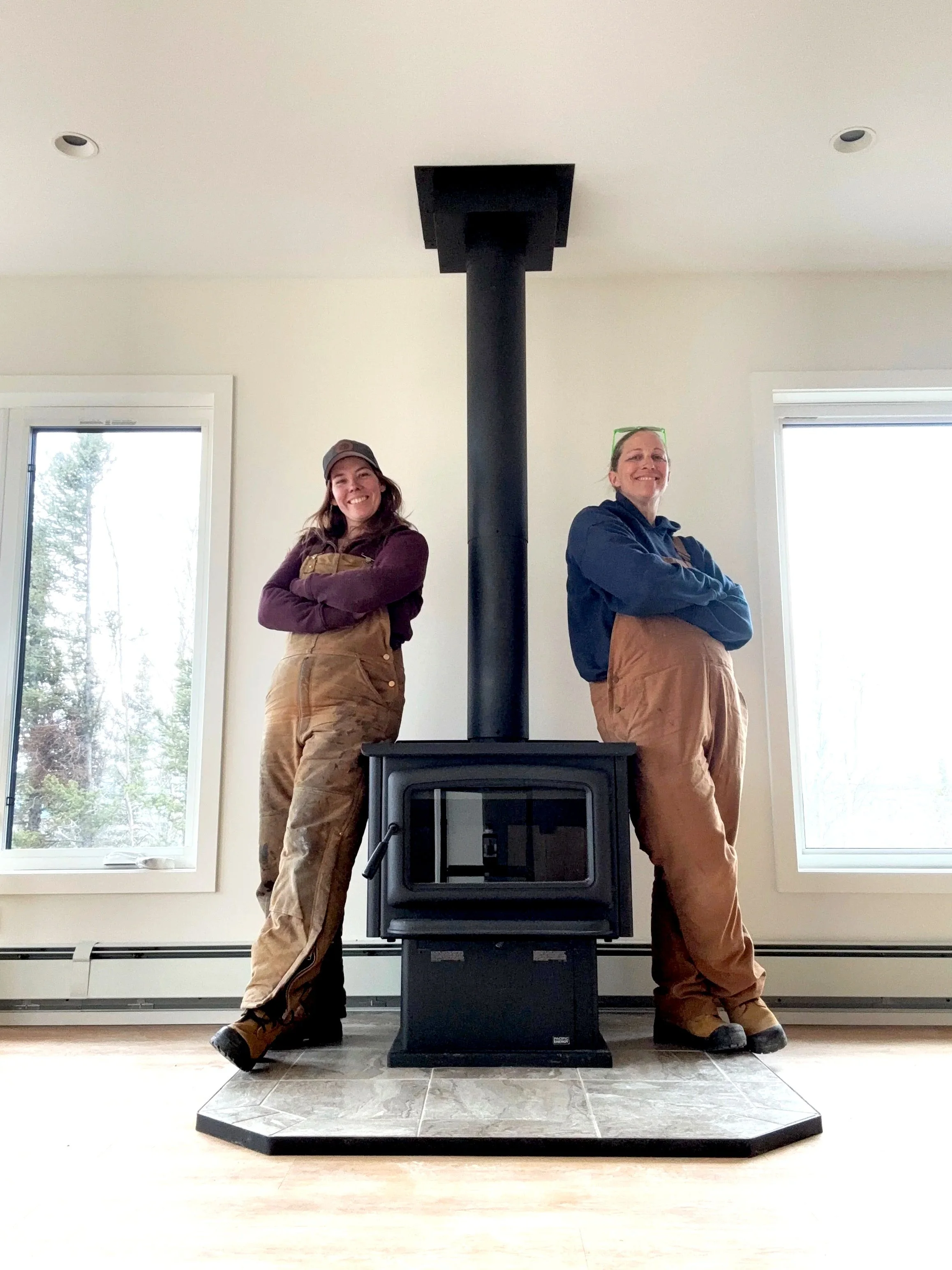 Two women standing on either side of a wood stove, smiling with arms crossed, inside a bright room with large windows.