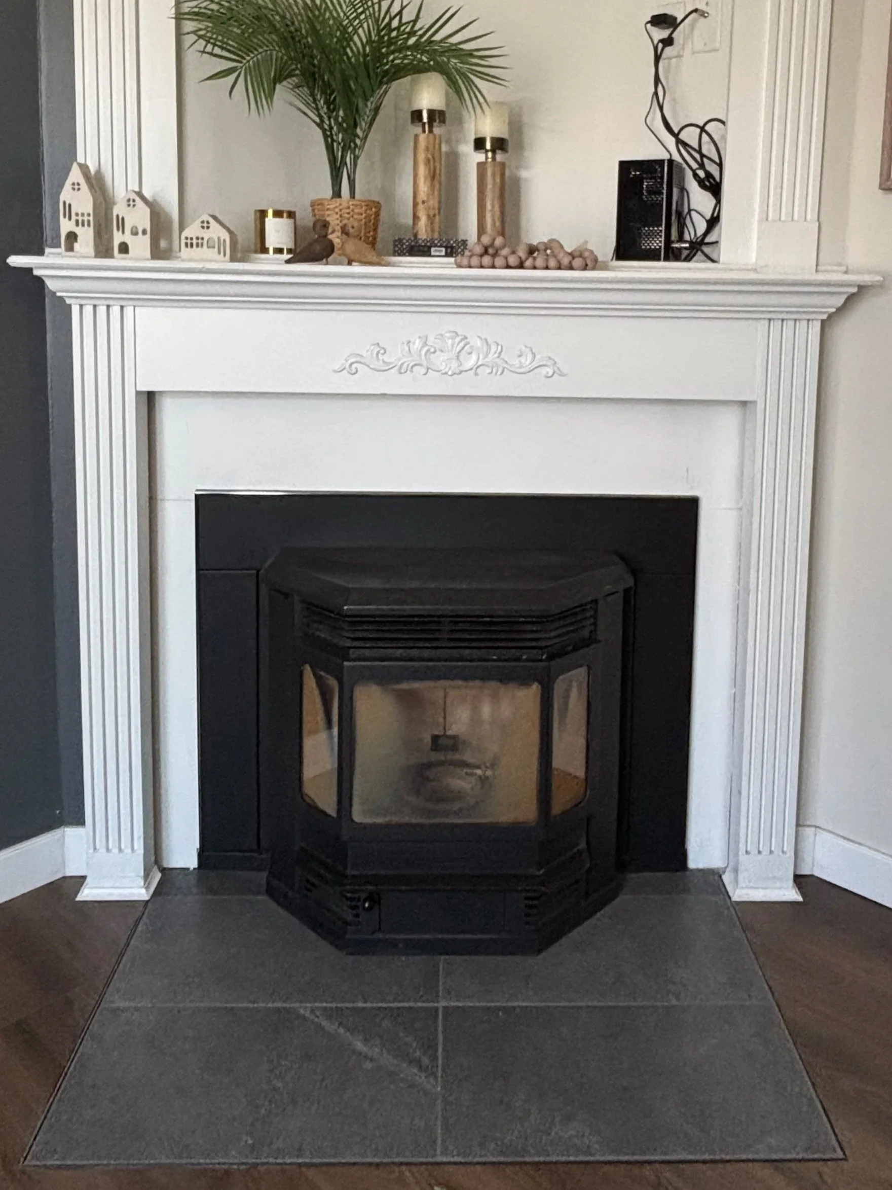 Decorative fireplace with a white mantel, featuring two small house-shaped ornaments, candles, a potted plant, and decorative items on top. A black stove insert is in the fireplace opening, and the hearth is tiled in dark gray.