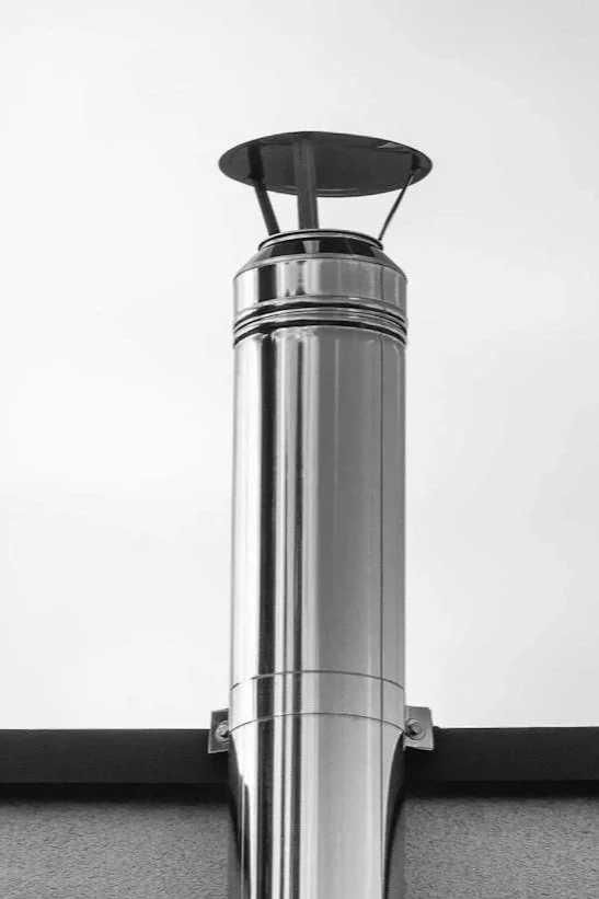 A black and white photo of a shiny metal industrial chimney or exhaust pipe extending vertically from a building, with a circular cap or cover at the top.