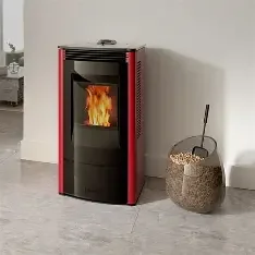 Modern pellet stove with a fire burning inside, positioned on a light-colored floor against a white wall. A decorative basket with a handle and a stick sits next to it.