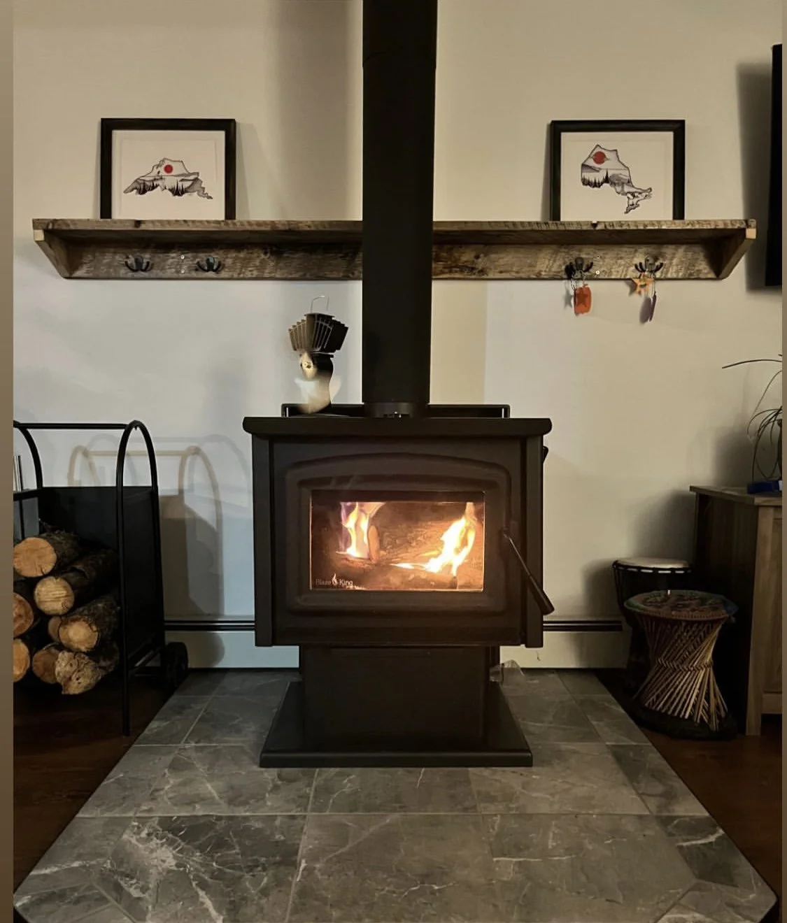 A living room with a fireplace with a fire burning inside, a black chimney pipe, a wooden mantle with framed wall art of a mountain and red sun, and decorative hooks with hanging ornaments. To the left, there's a log holder with stacked logs, and to the right, a small wooden table with a plant.