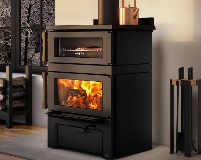 Black wood-burning stove with a glass front showing a fire, in a cozy indoor setting with a window and stacked firewood nearby.