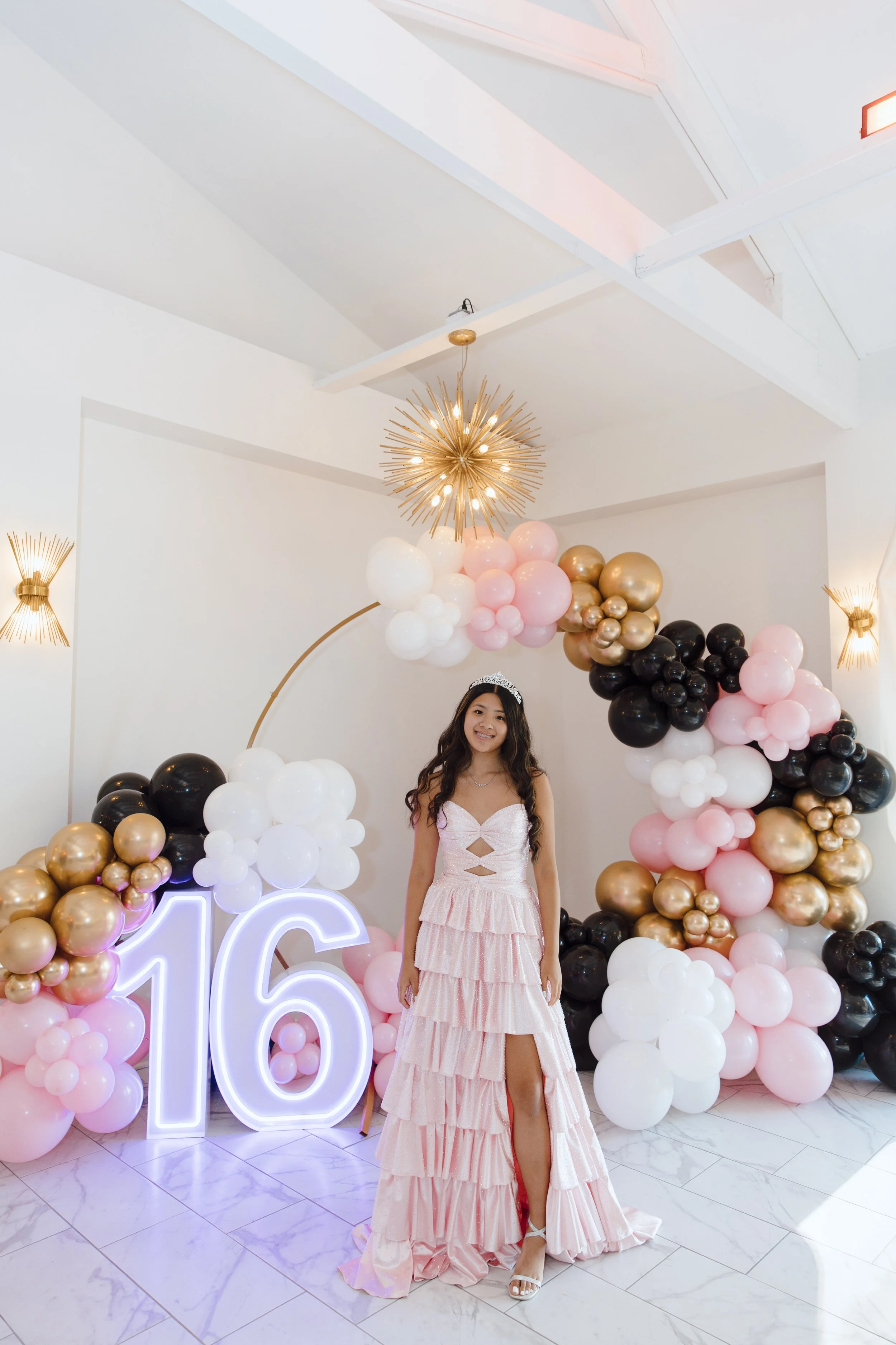 A young woman in a pink dress smiling at a birthday celebration, standing in front of balloon decorations and a neon number 16 sign.