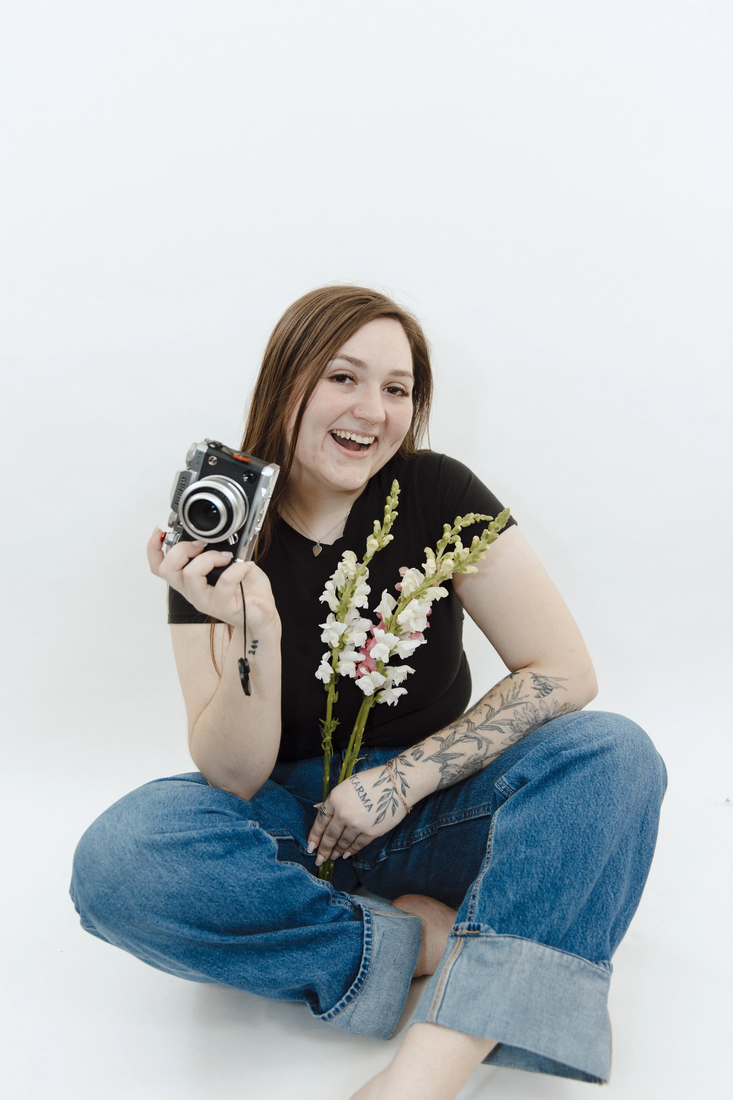 A young woman with brown hair sitting cross-legged on the floor, smiling, holding a camera in one hand, a bouquet of white flowers in the other, wearing a black t-shirt and blue jeans with tattoos visible on her left arm, against a plain white background.
