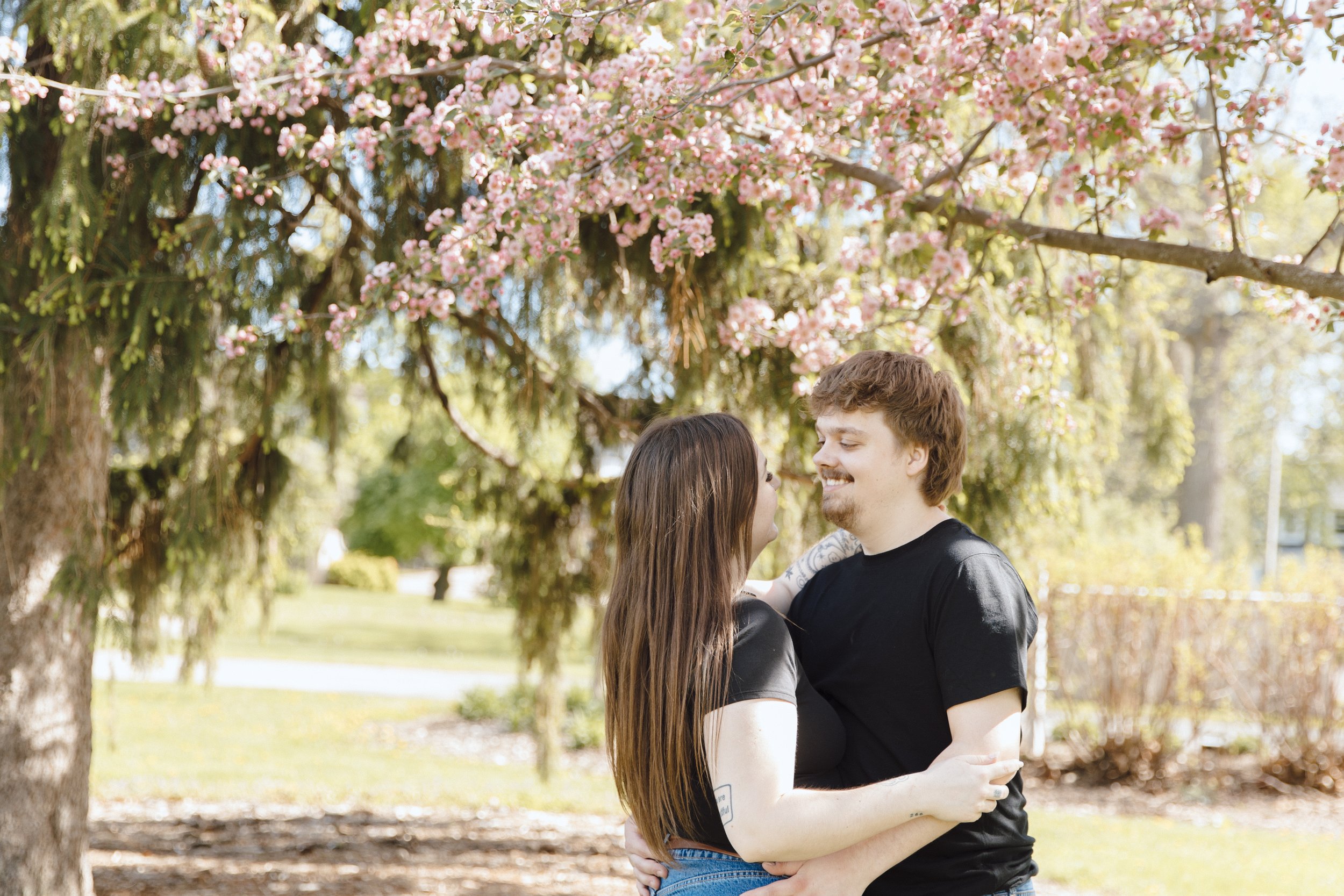A young couple embraces outdoors in front of a blooming tree with pink flowers, smiling at each other.