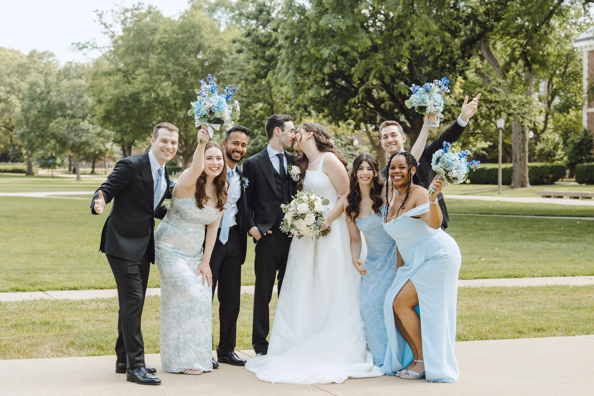 A bride and groom standing in the middle of a group of people who are wearing blue dresses, black suits with blue ties, and folding bouquets of flowers. Photo by Lumen Photography Studios, based in DeKalb, Illinois.