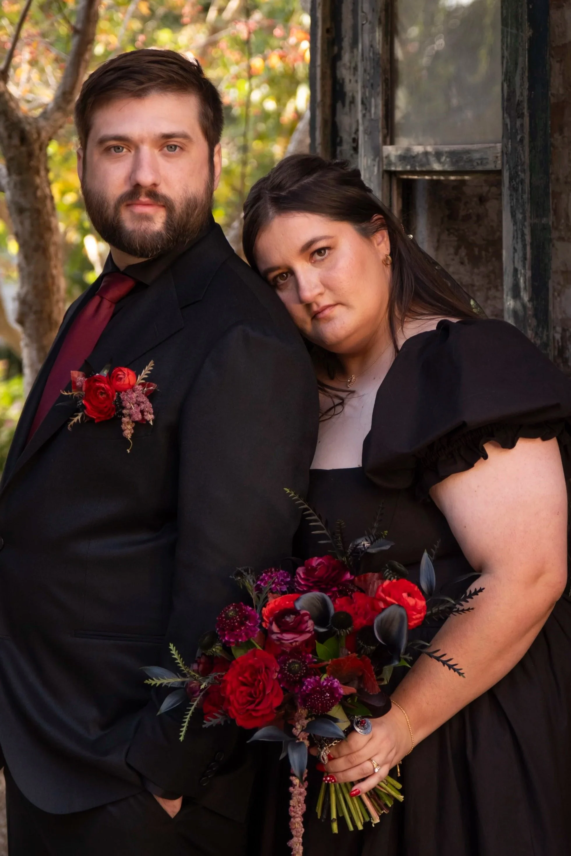 A man and woman pose outdoors, the woman resting her head on the man's shoulder. The man is dressed in a black suit with a red tie and a flower boutonniere. The woman wears a black dress with puffed short sleeves and holds a large bouquet of red and purple flowers. They are standing next to an old, rustic wooden structure with trees in the background.