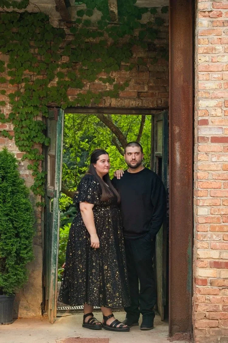 A woman and a man standing in front of an open doorway surrounded by brick walls, with green foliage visible outside.