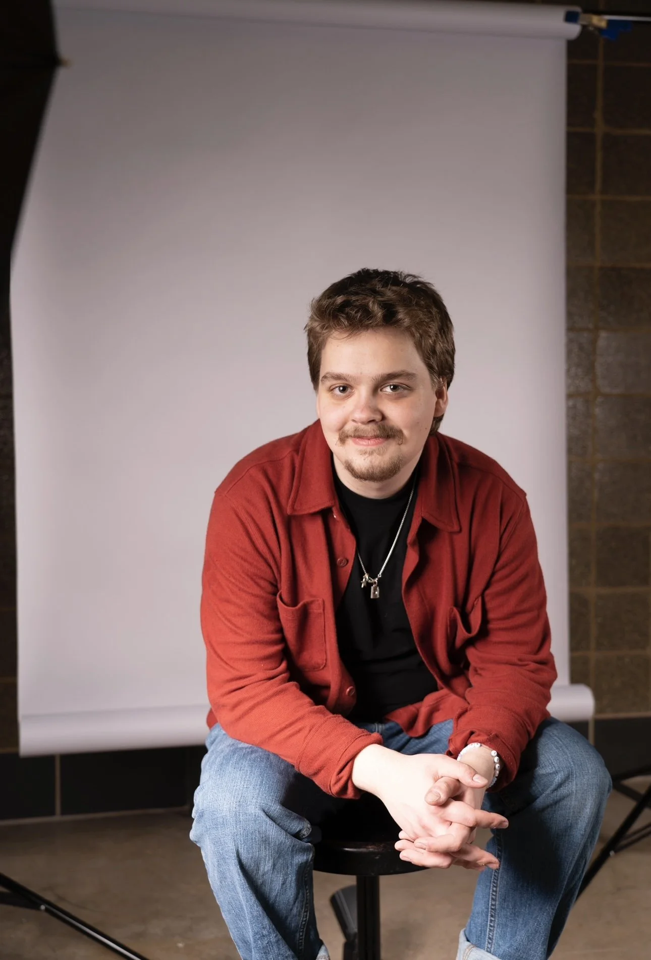 A man with brown hair, a goatee, and mustache, wearing a red shirt over a black T-shirt, sitting on a stool in front of a white backdrop, with his hands clasped.