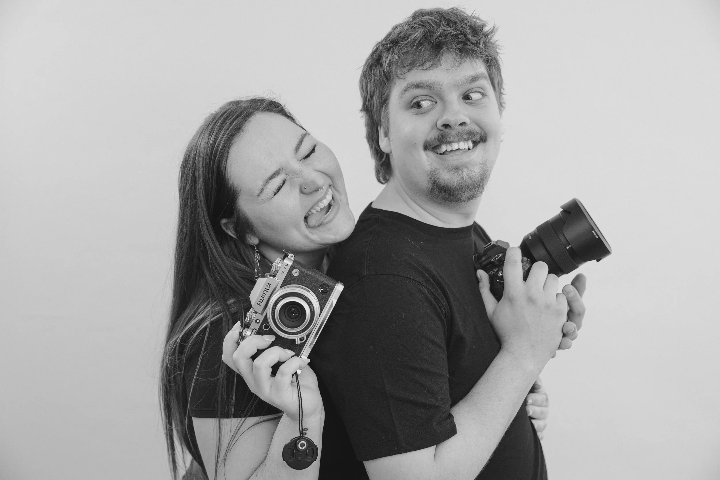 Black and white photo of a smiling woman with long hair and a nose ring, holding a camera, hugging a man with short hair, a content expression, and holding a camera, against a plain white background.